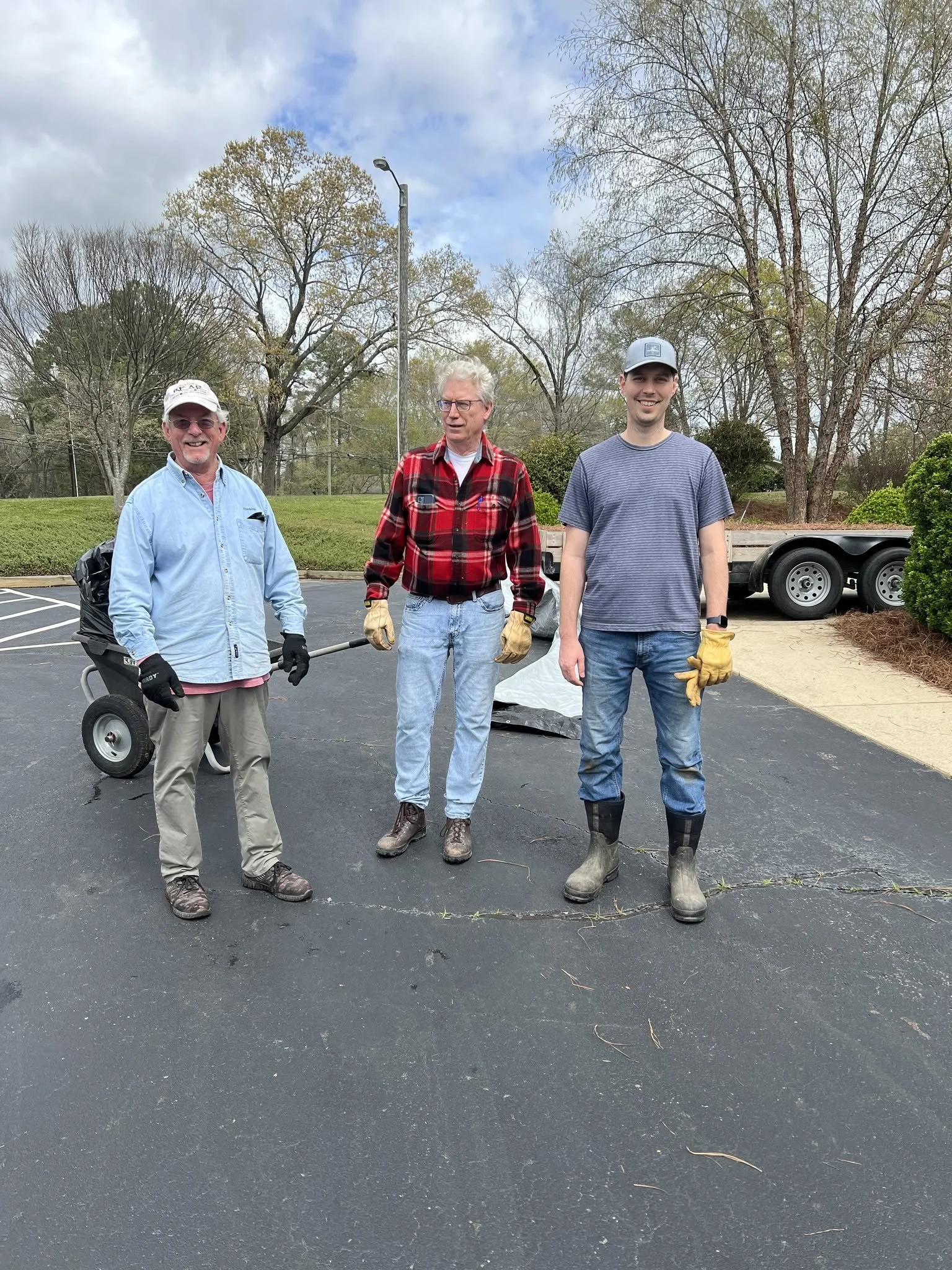 Three men standing on a paved surface outdoors, smiling at the camera. The man on the left is wearing glasses, a light blue shirt, and khaki pants, holding a tool. The man in the middle has gray hair, glasses, a red plaid shirt, and jeans. The man on