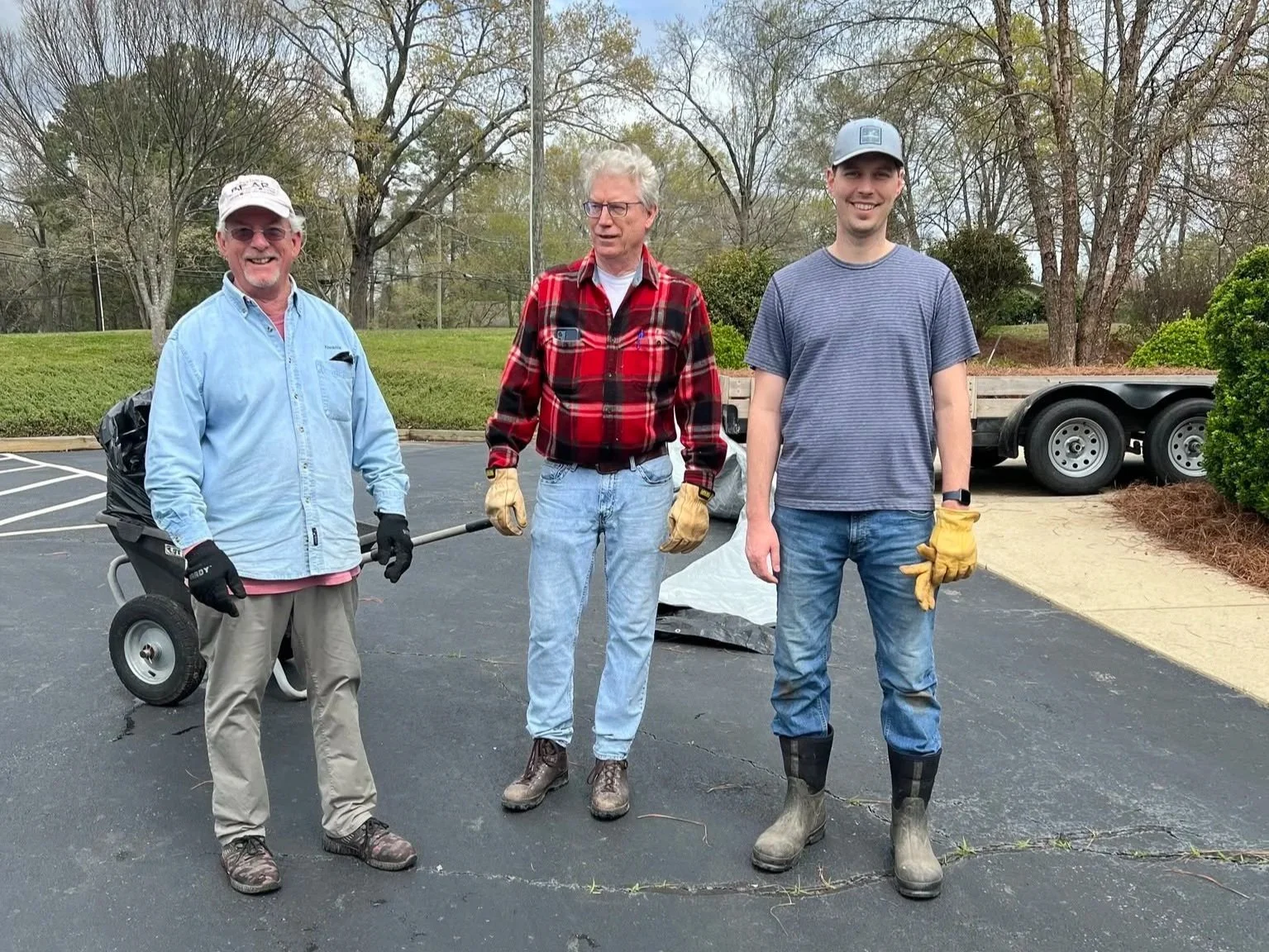 Three men standing outdoors in a parking lot, wearing work gloves and casual clothes. They appear to be involved in a cleanup or maintenance activity, with a cart and a trailer visible in the background, along with trees and bushes.