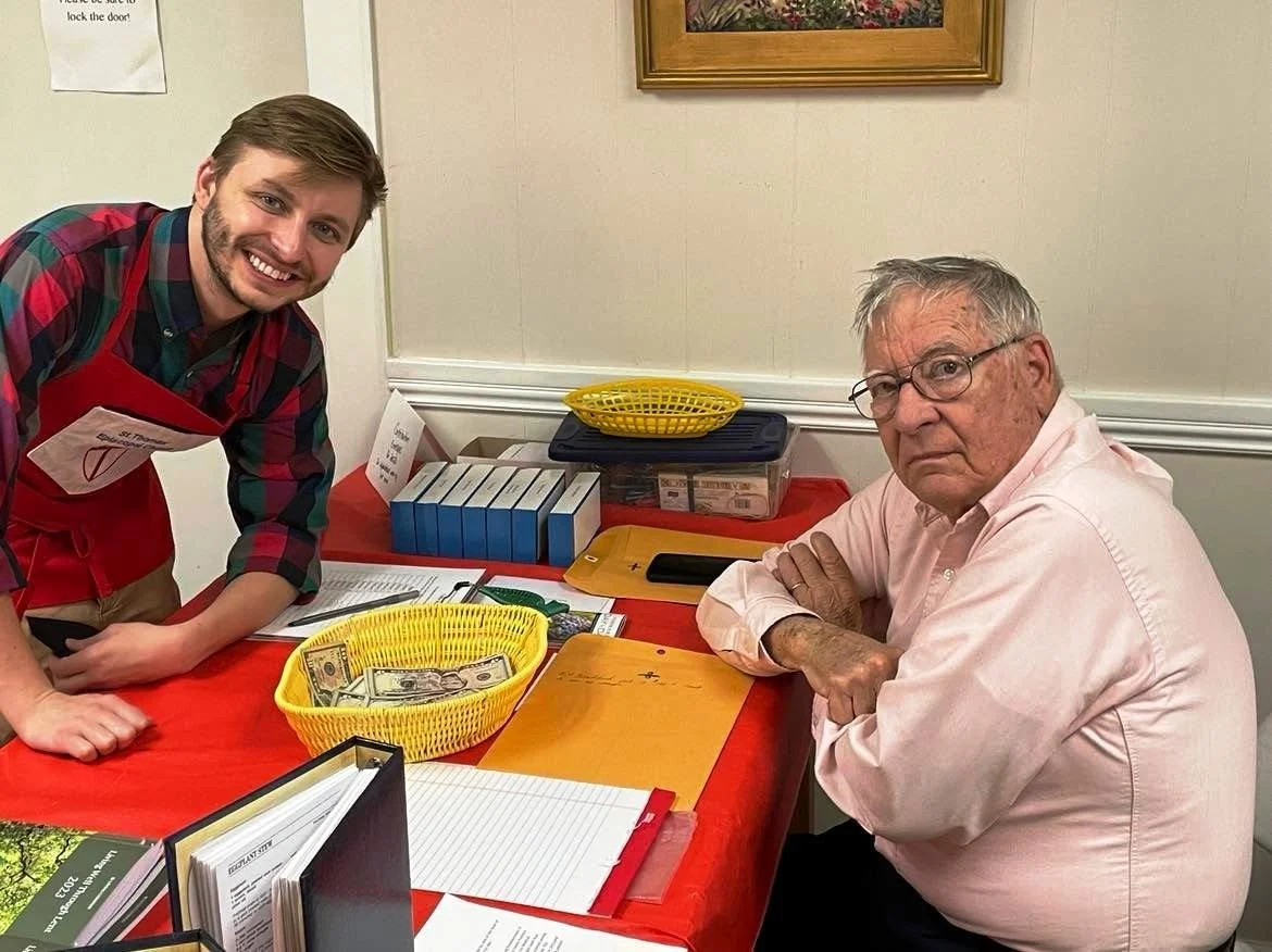 A young man in a red apron and plaid shirt smiling at a camera standing beside an older man with glasses in a pink shirt sitting at a table with cash in a yellow basket. The table has papers, binders, and other office supplies, in a room with framed 