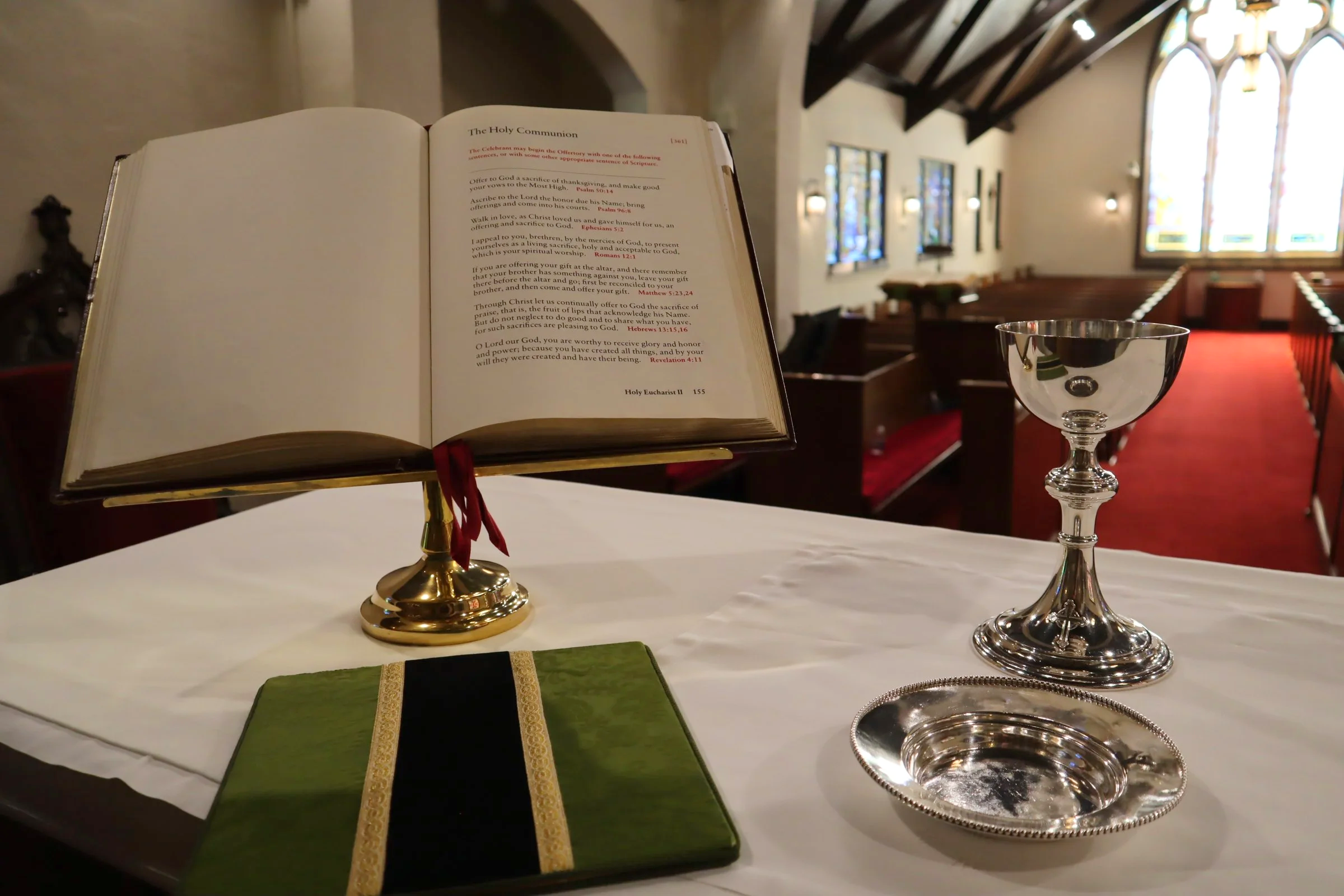 Open Bible on a stand, communion chalice to the right, green cloth with gold trim, and a silver tray on a white altar cloth inside a church with stained glass windows and wooden pews.