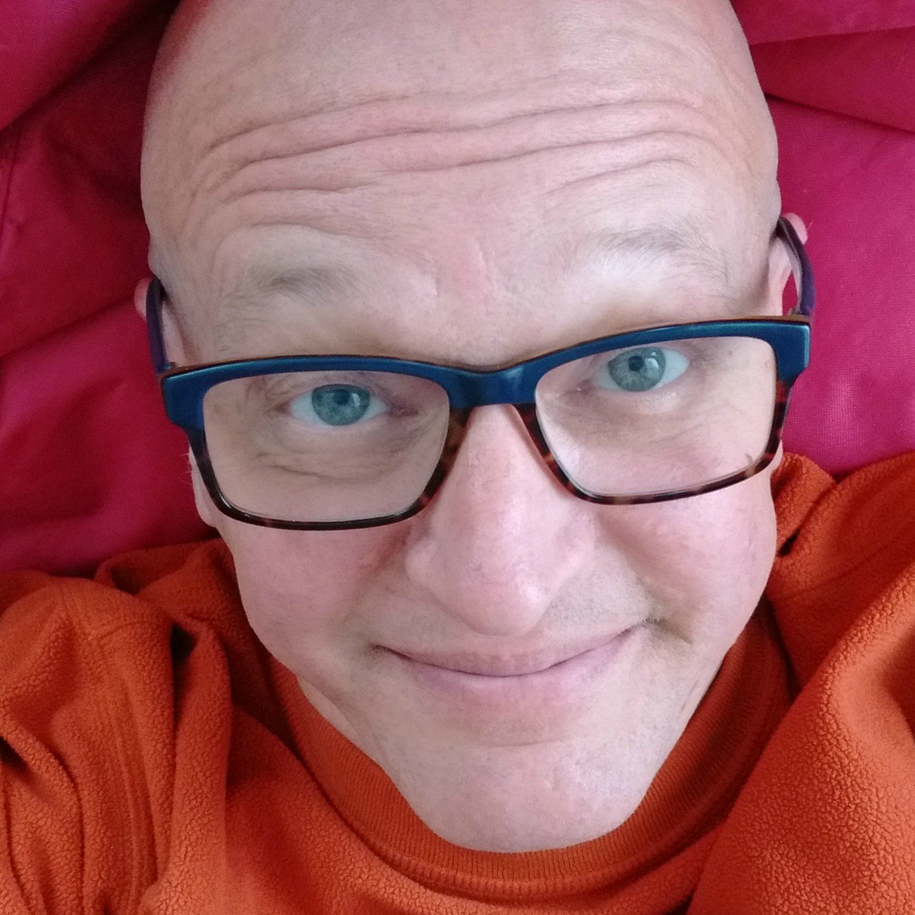 A person with light skin, glasses, and blue eyes, smiling while lying down on a pink surface, wearing an orange hoodie.
