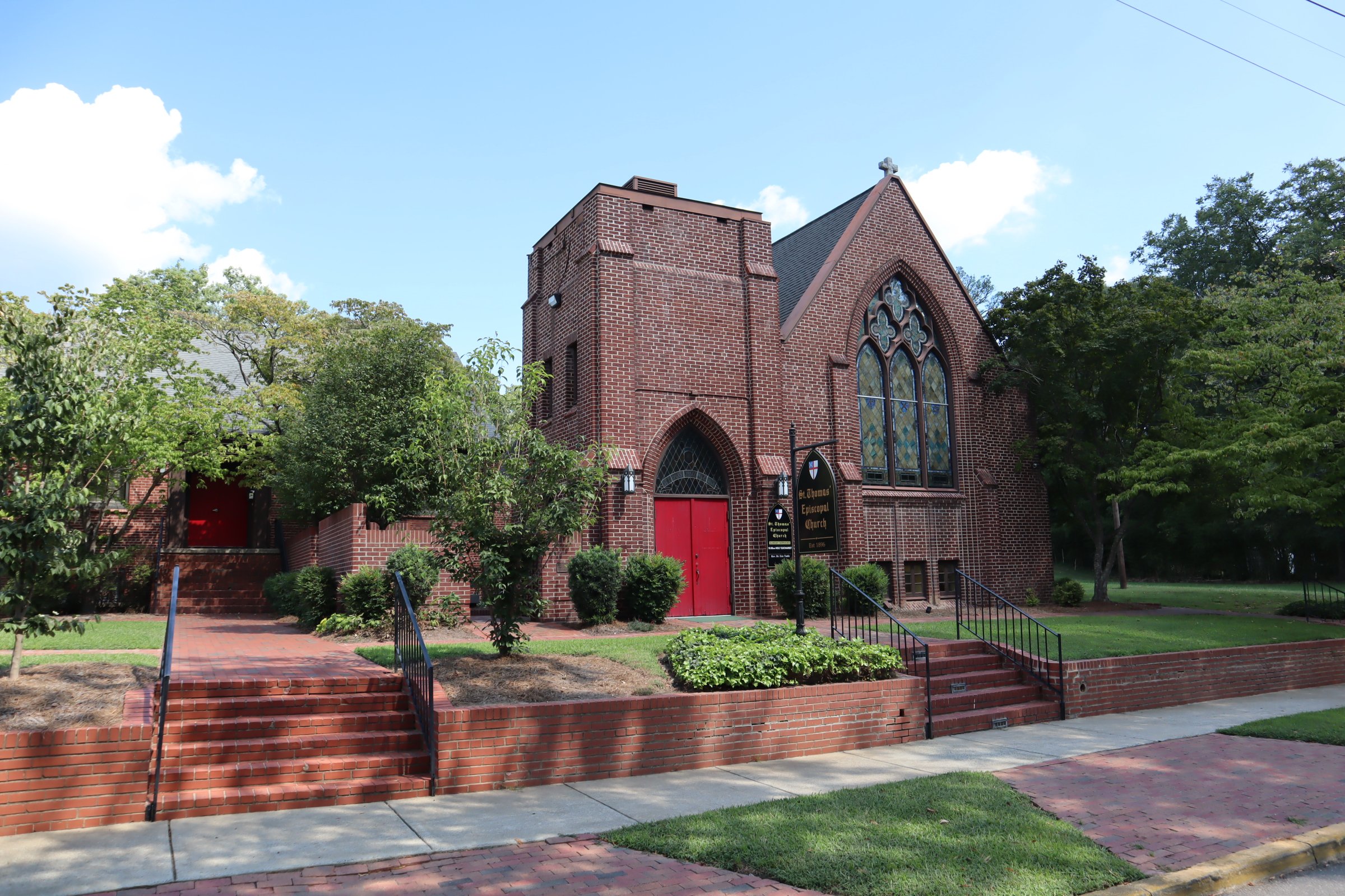 Traditional and historic brick church with pointed roof and stained glass windows, surrounded by trees, with a sign that reads 'St. Thomas Episcopal Church.'