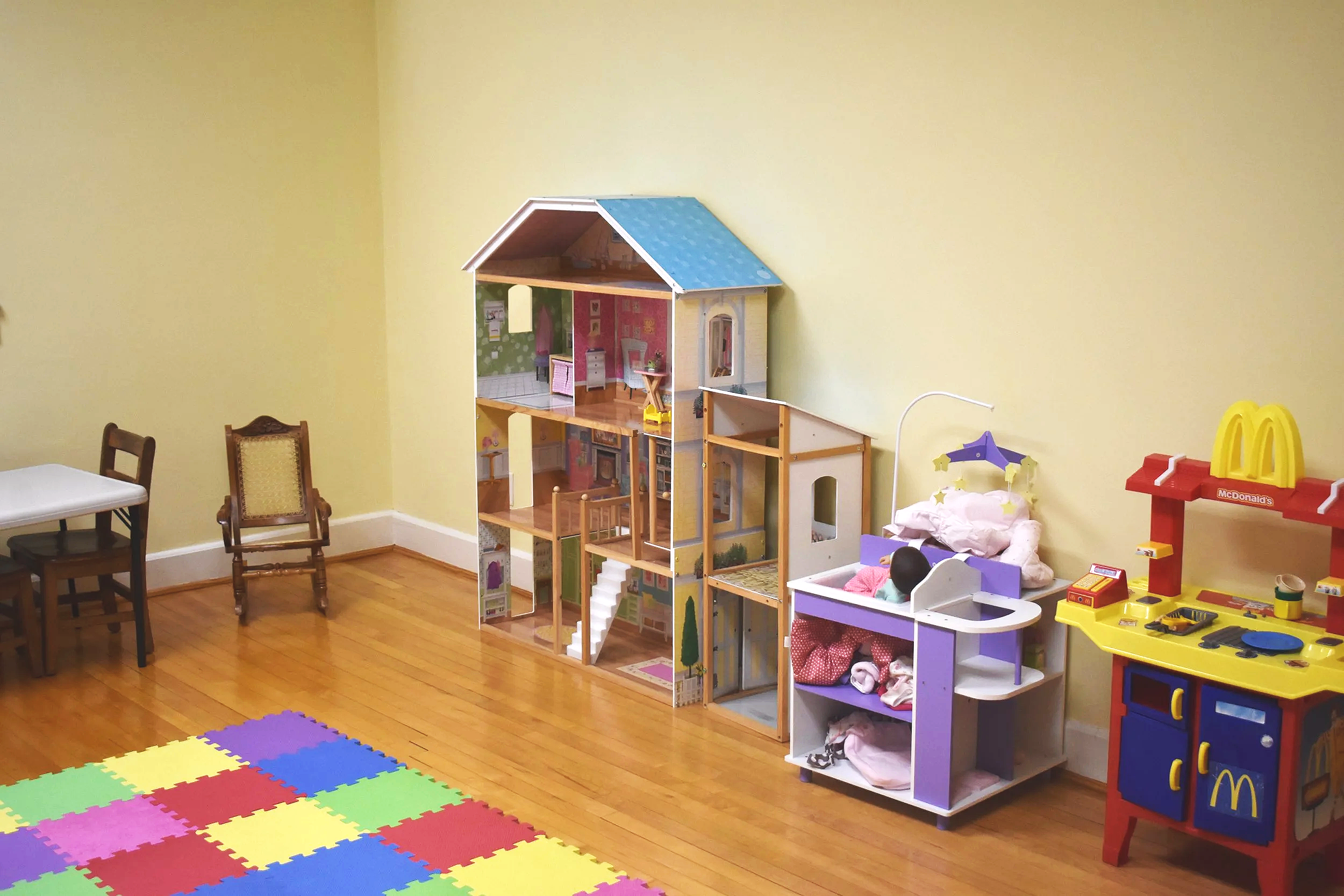 Colorful children's playroom with a dollhouse, toy kitchen, and chairs against a yellow wall, and foam puzzle mats on the wooden floor.