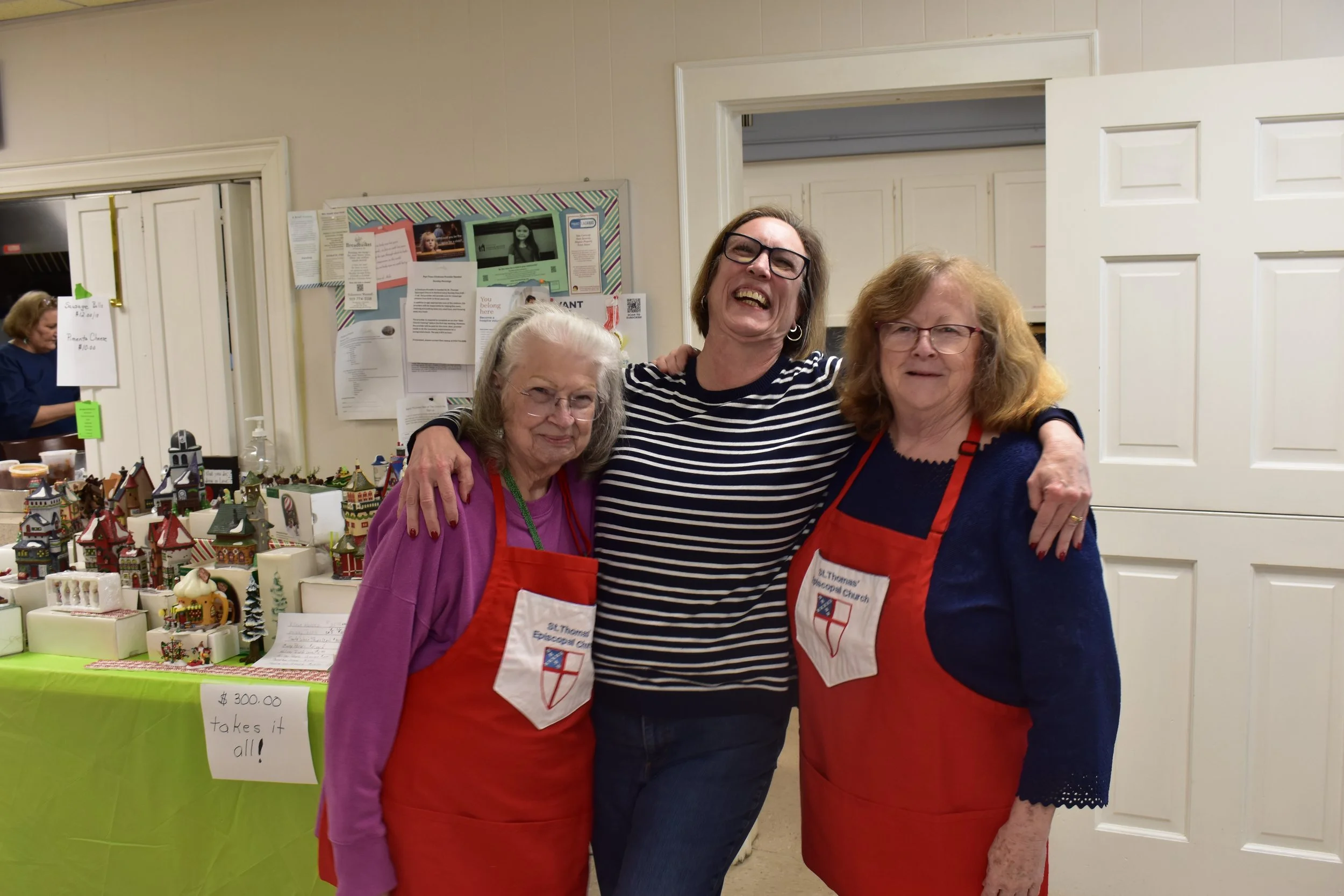 Three women wearing red aprons visiting a craft fair booth with miniature holiday houses. They are smiling and standing close together, with a woman in the background at a different booth.