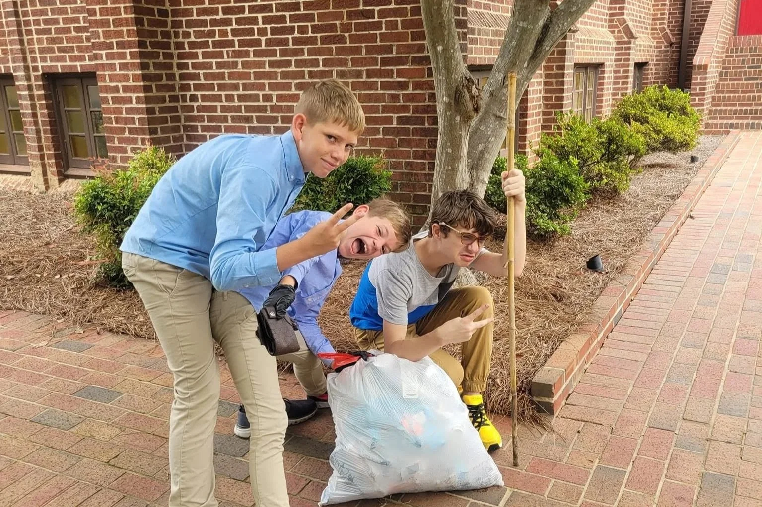 Three boys cleaning up trash outside near a brick building, holding trash bags and a trash picker.
