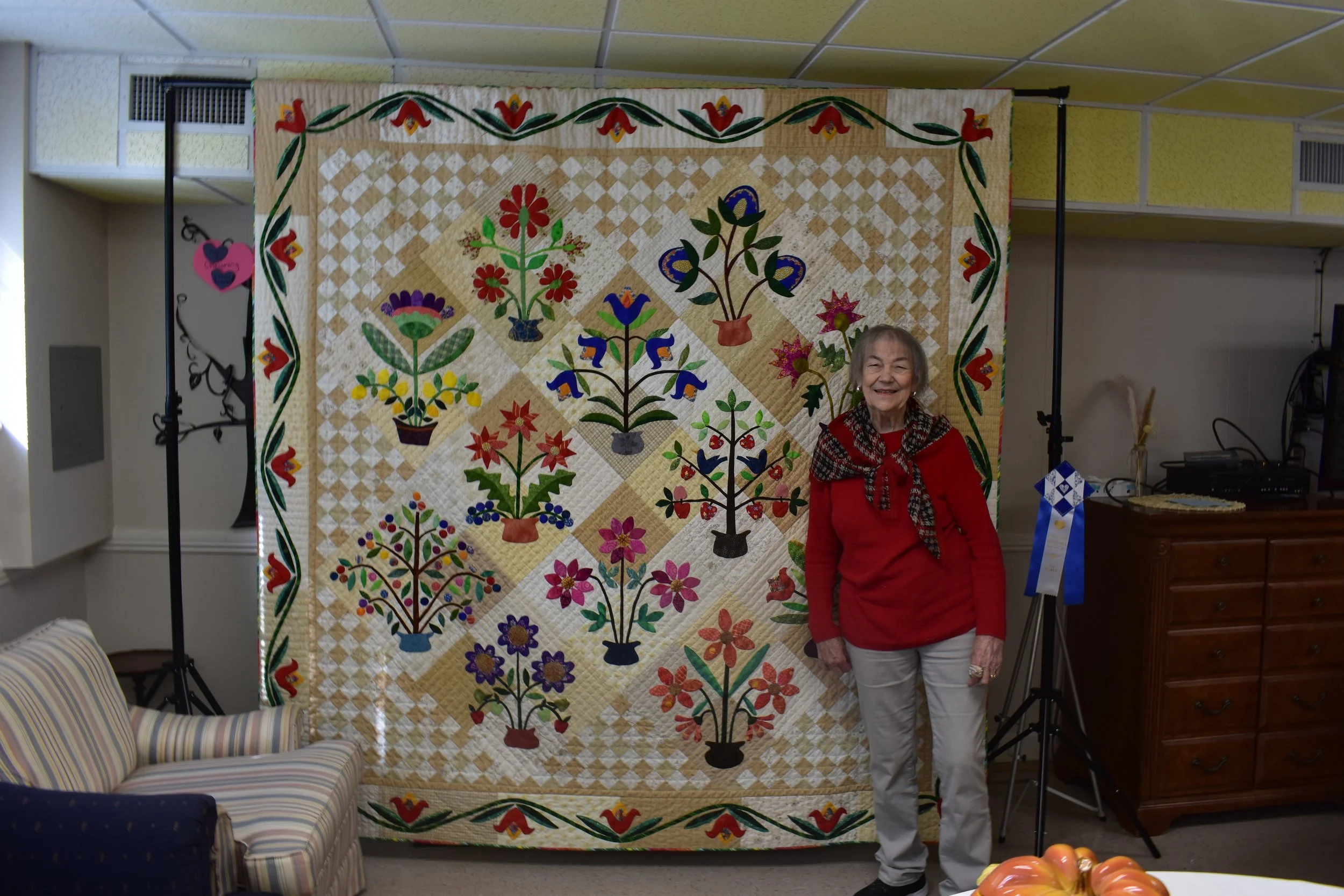 An elderly woman with gray hair smiling, wearing a red sweater with a black and white scarf, standing indoors in front of a large colorful quilt featuring various flower and plant motifs, with a floral border. The background includes a beige wall, a 