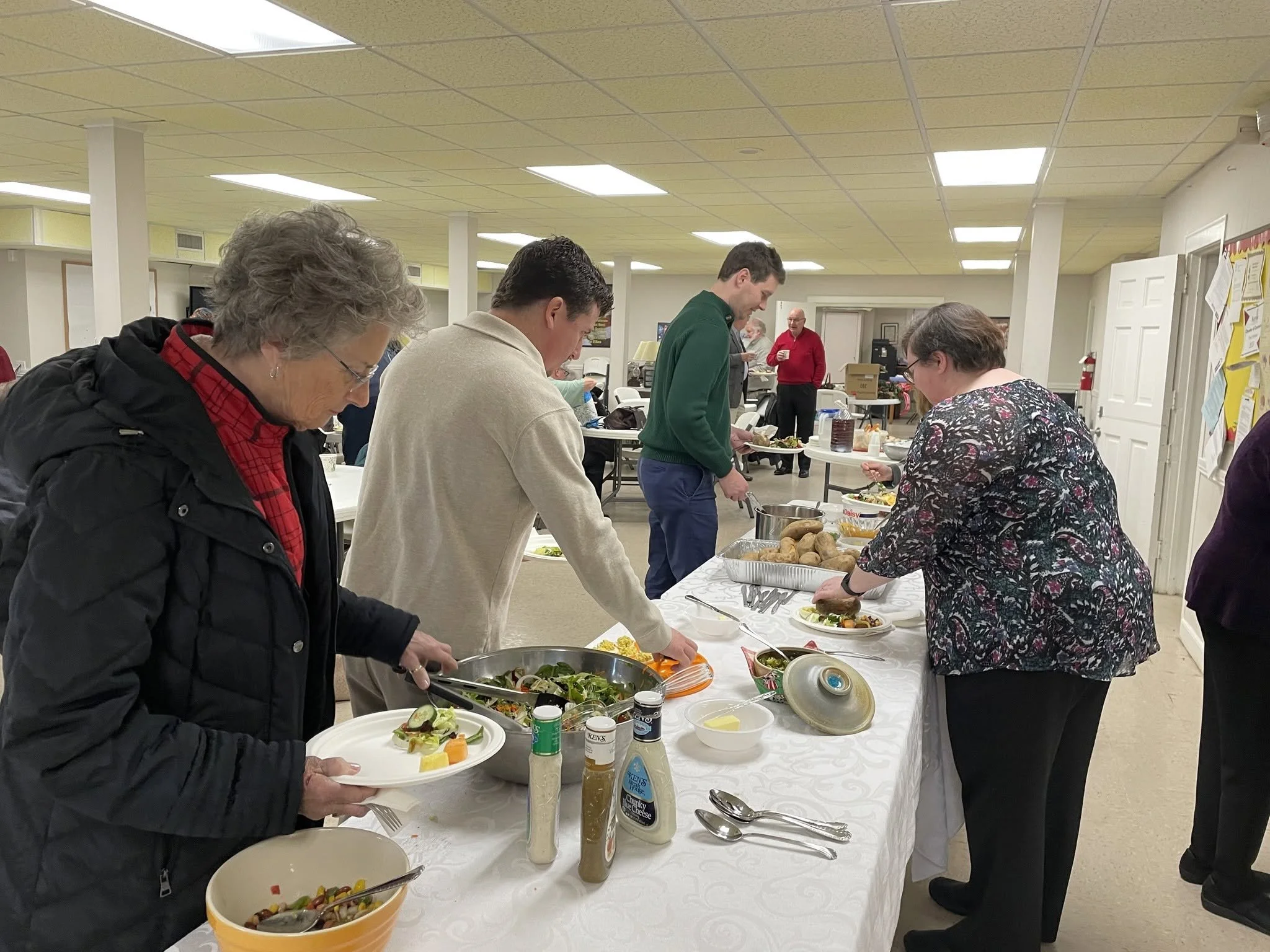 People standing in line at a buffet table with various salads and condiments in a community center or cafeteria setting.