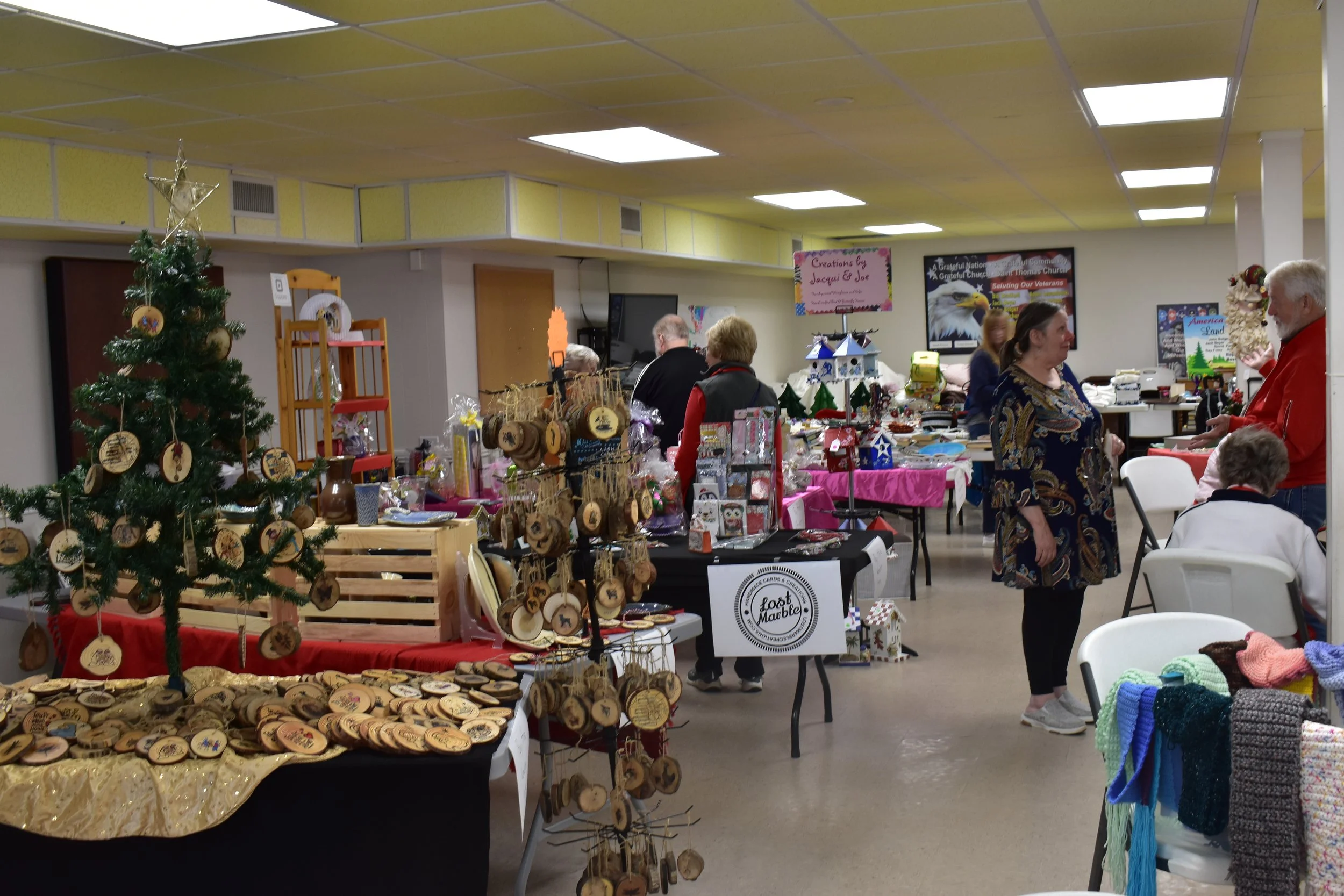 Indoor craft fair with tables displaying wooden ornaments, handmade crafts, and knitted items. Christmas decorations, a small Christmas tree, and vendors engaging with customers are visible.
