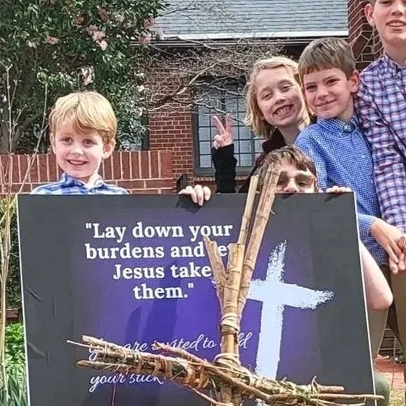 Group of children standing outdoors holding a sign with a religious message, a cross, and some sticks in front.