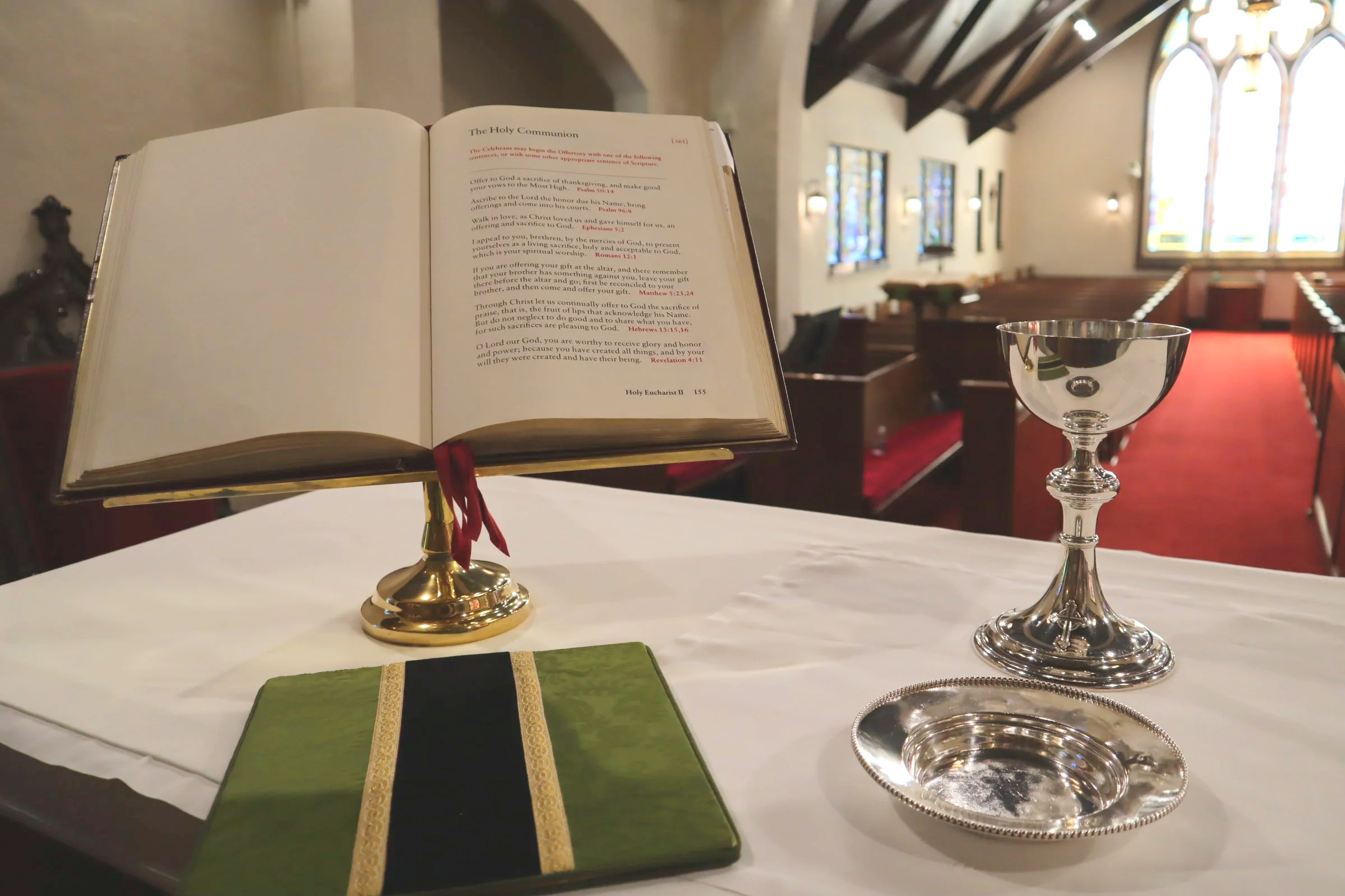 An open Bible on a altar or table inside a church, with a chalice and a green-covered book nearby. The background shows church pews, stained glass windows, and wooden ceiling beams.