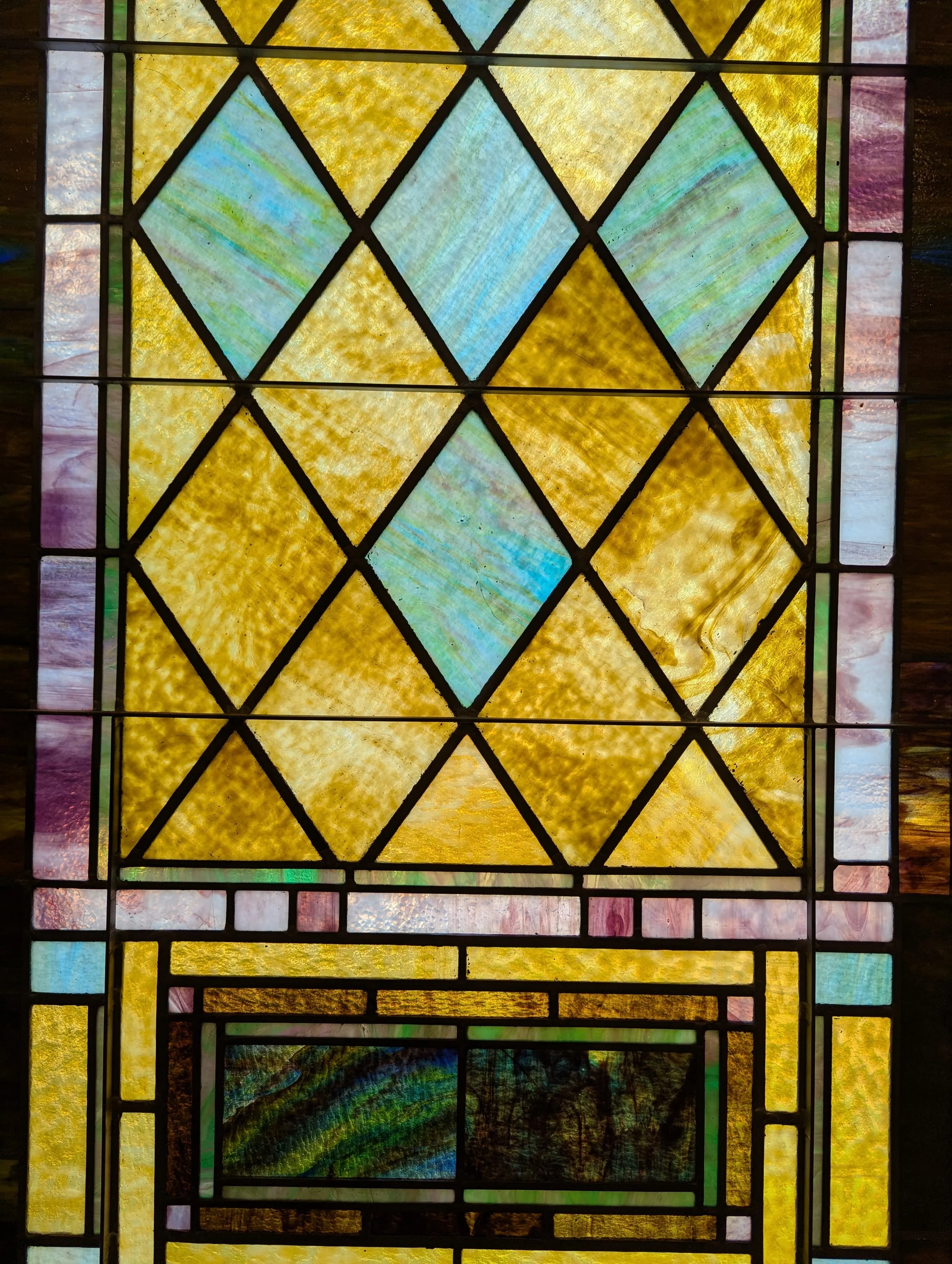 Colorful stained glass window with geometric patterns, including diamonds and rectangles, featuring yellow, blue, pink, and green hues.