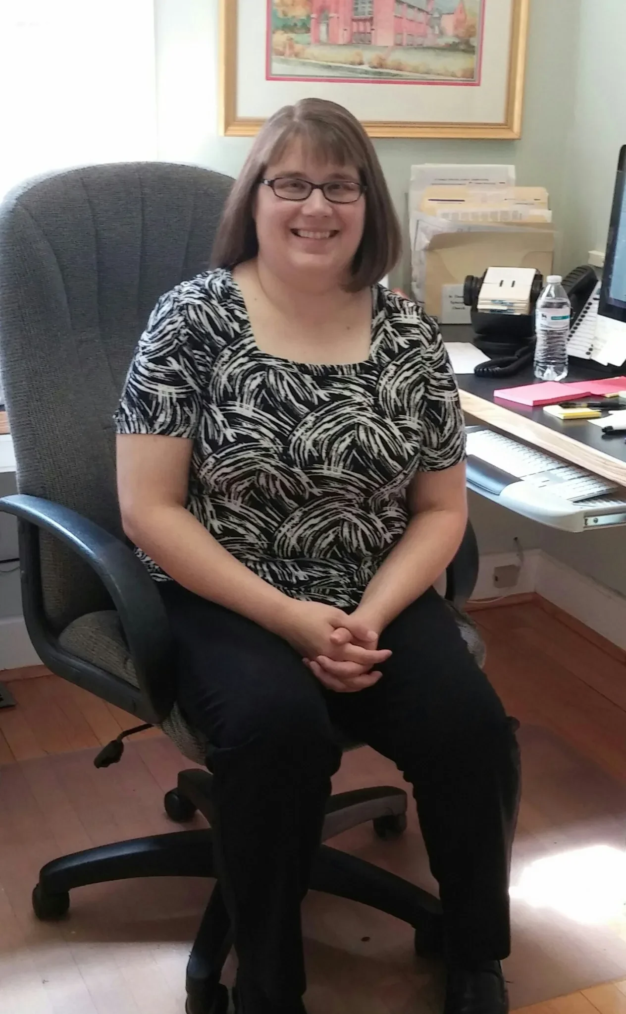A woman with glasses and shoulder-length brown hair sits in an office chair, smiling. She is wearing a black and white patterned top and black pants. Behind her is a desk with papers, a computer, a water bottle, and office supplies. There is a framed picture on the wall above the desk.