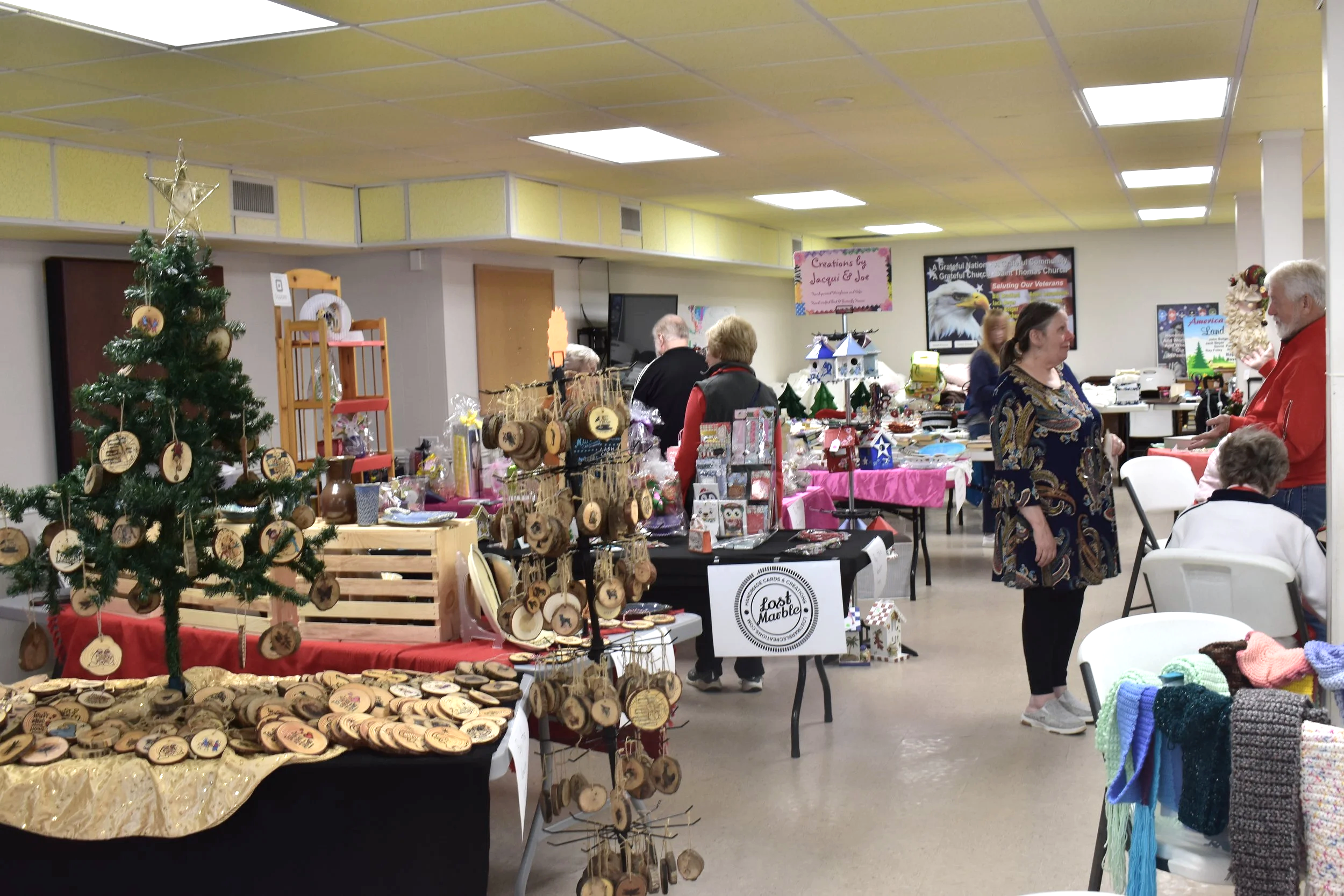Indoor holiday craft fair with tables displaying handmade items, Christmas decorations, and people browsing and talking.