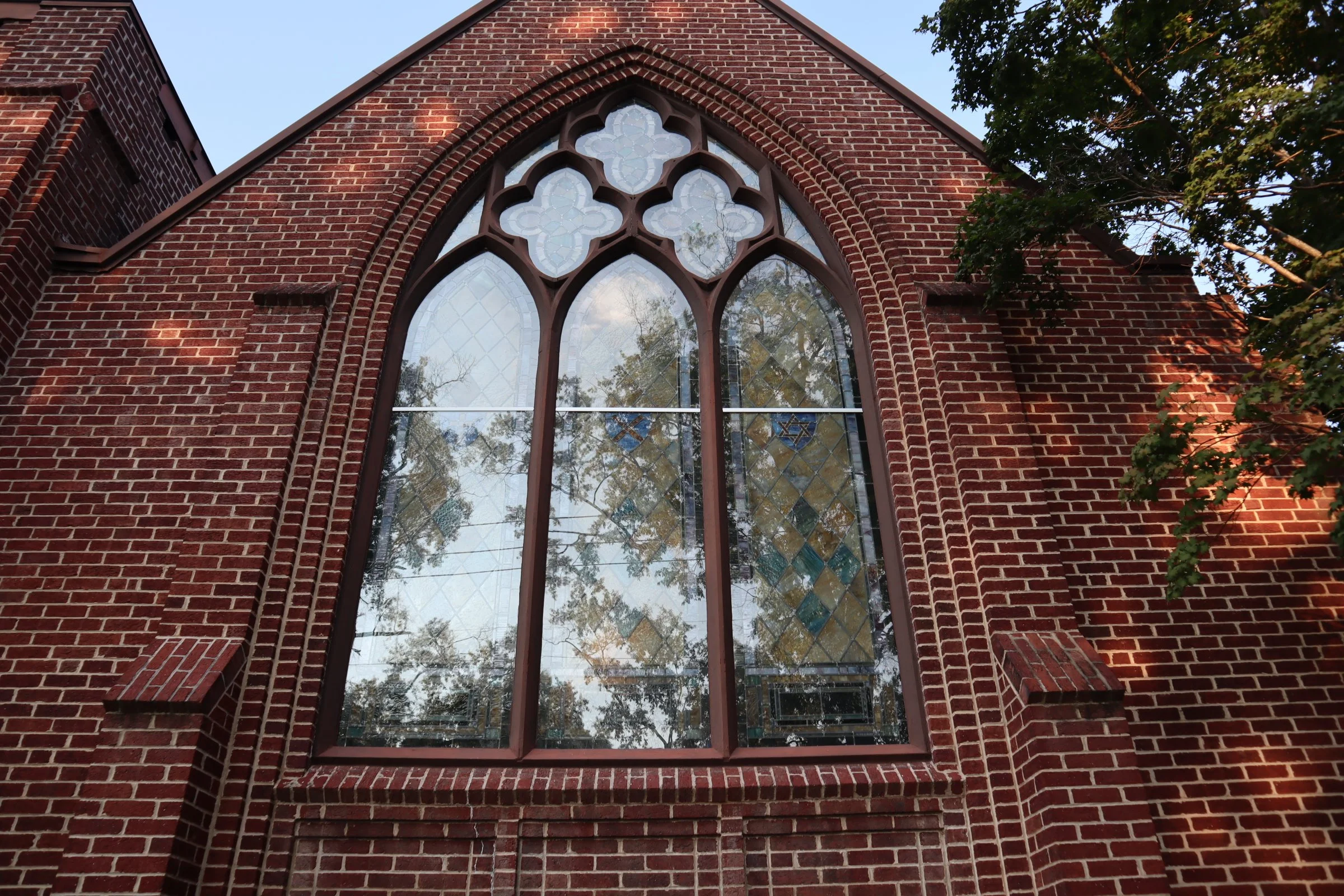 A close-up view of the facade of a red brick church with a large arched stained glass window, partially covered by tree branches.
