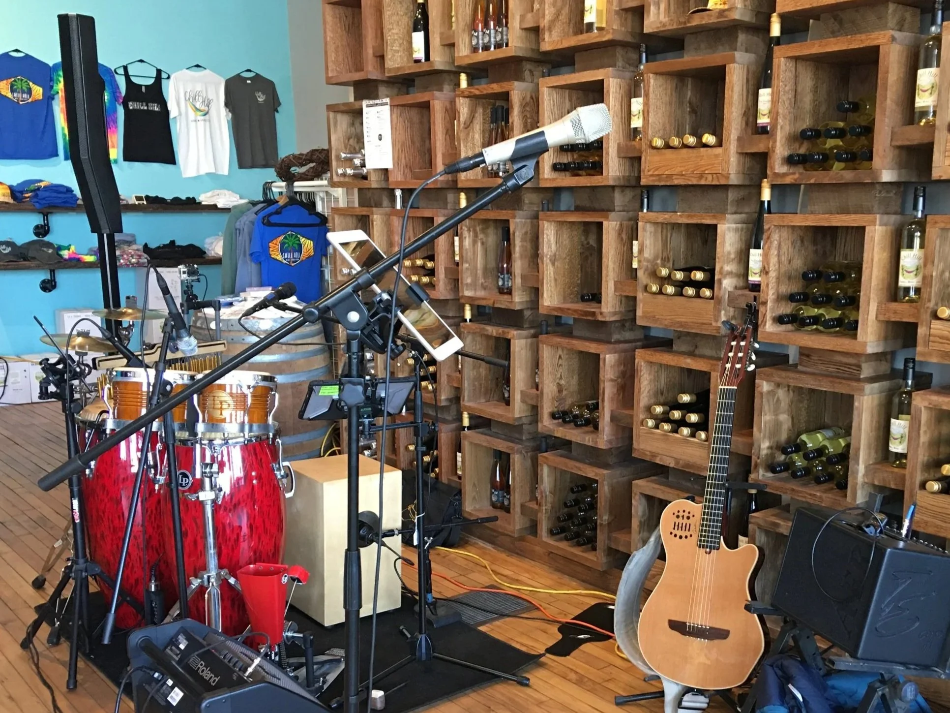 Coastal Vibes acoustic music setup and wine shop interior with musical instruments, microphones, wine bottles on wooden shelves, and T-shirts on the wall.