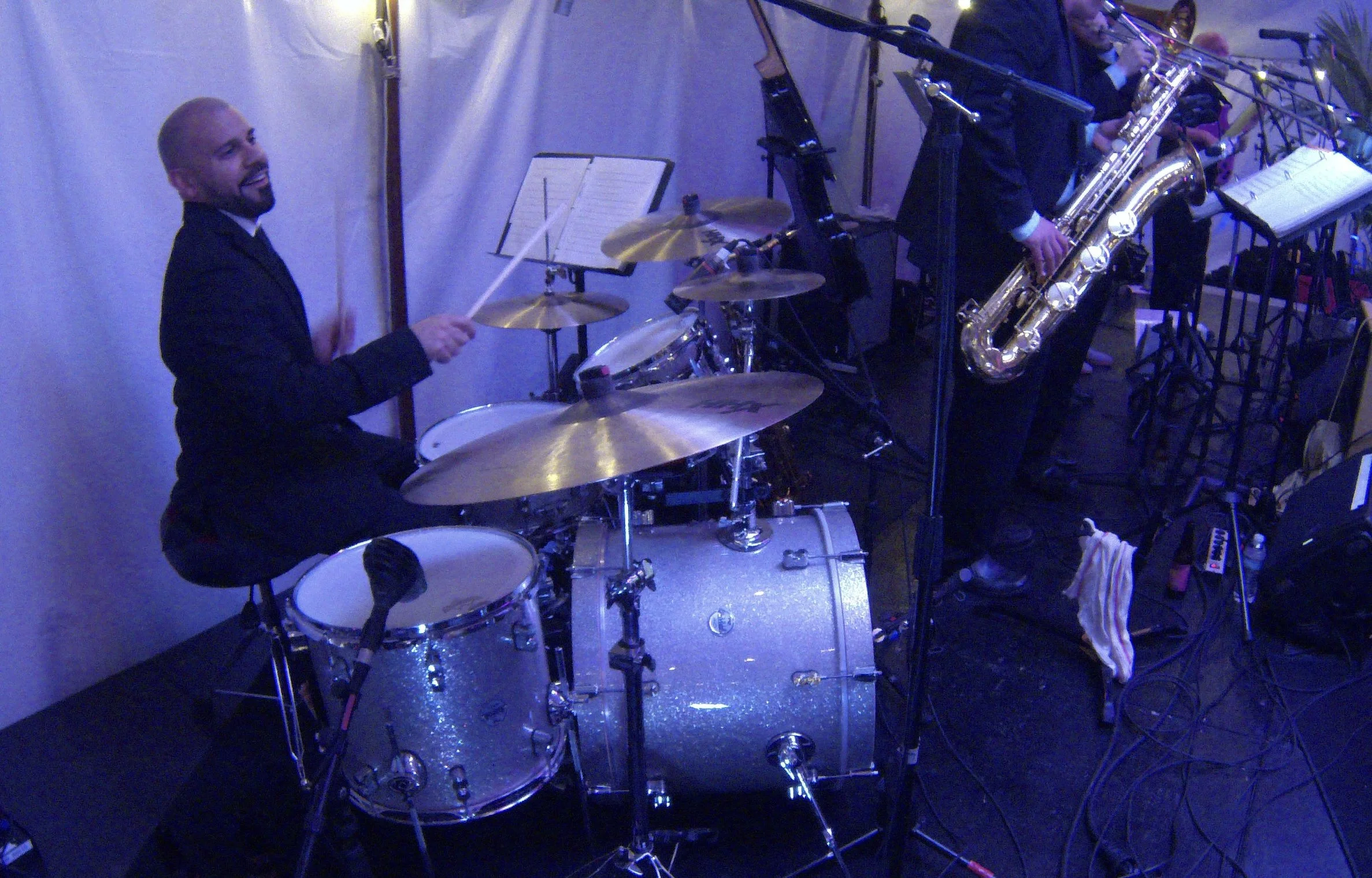 James Rodriguez on drums with a corporate band performing on stage, dressed in formal black attire. The stage is lit with blue lighting.