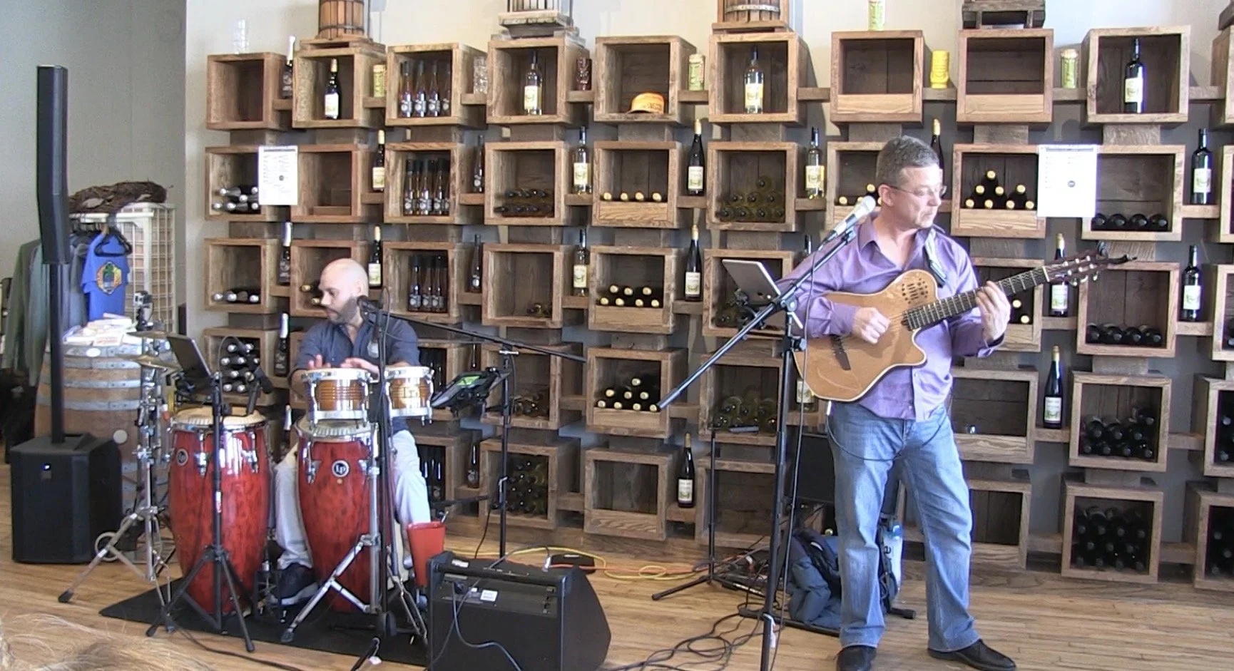James Rodriguez & Dale Neville perform in a wine cellar. One plays conga drums, and the other plays an acoustic guitar. The background consists of wooden wine bottle storage boxes mounted on the wall.