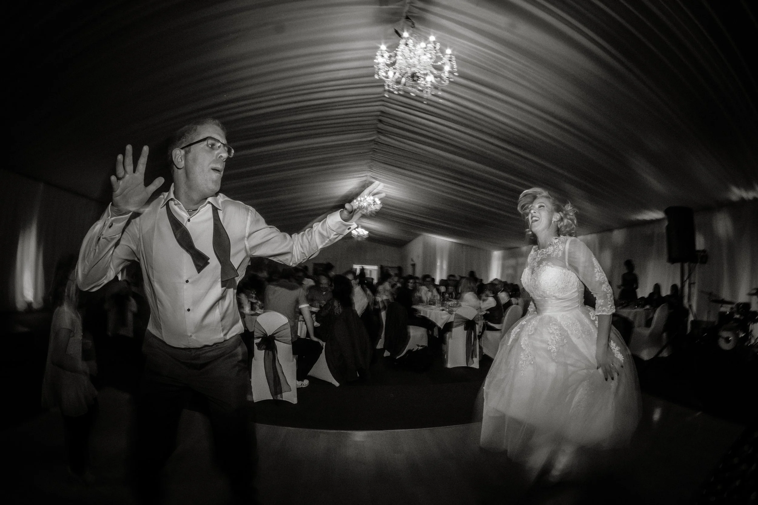 A bride in a floral lace wedding dress dancing with a groom in a tuxedo at a wedding reception, with guests seated at tables in the background, under a wooden ceiling and a chandelier.