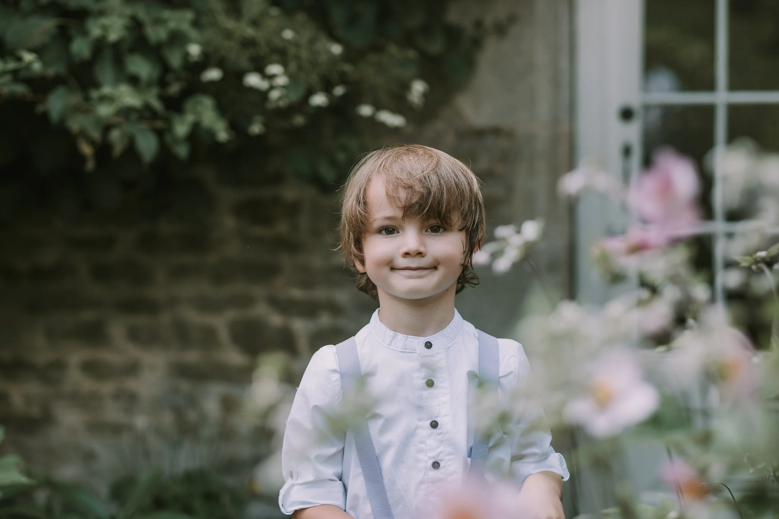 Young boy with light brown hair smiling, wearing a white shirt with gray suspenders, standing outdoors in a garden area with bushes and flowers.