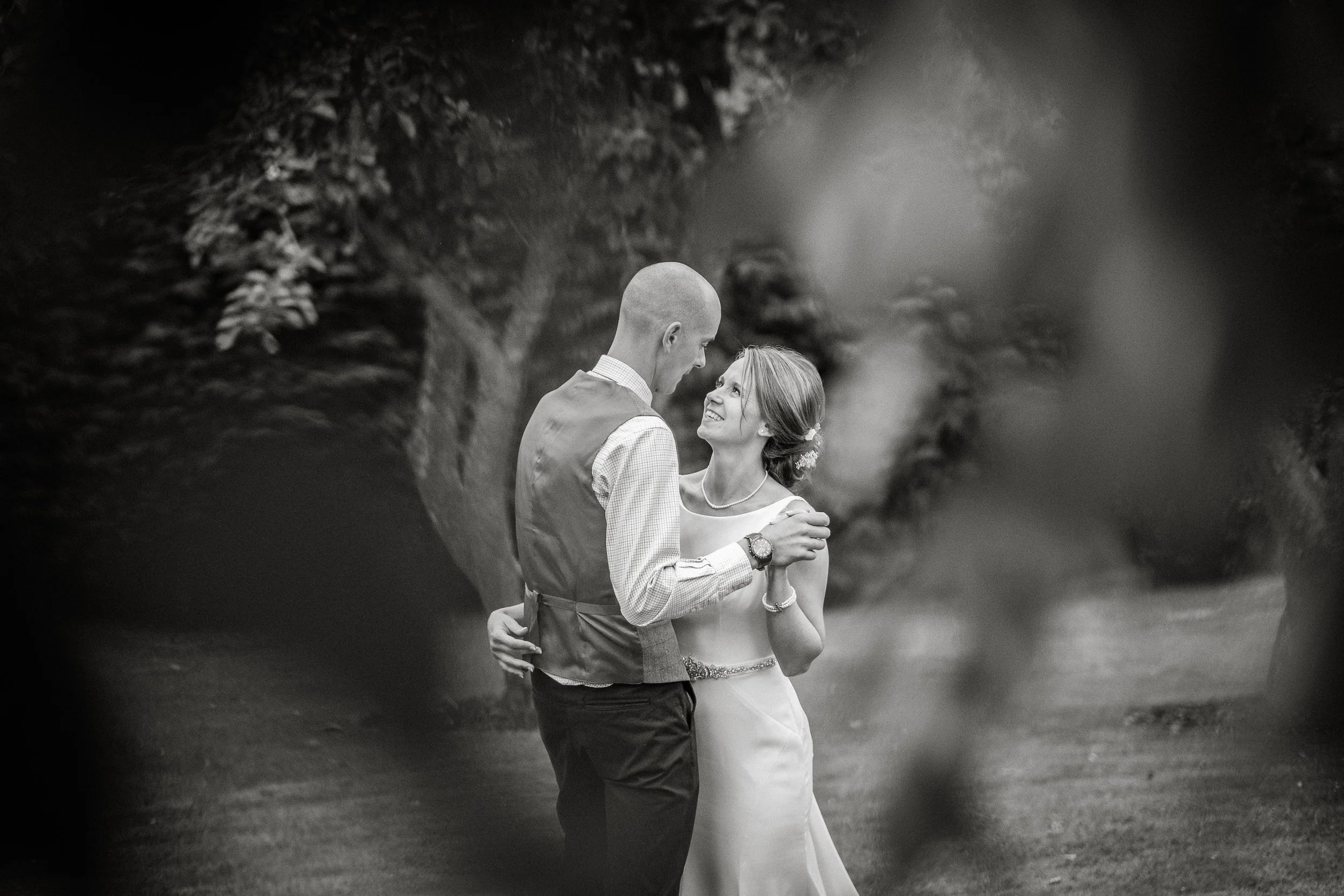 A couple, dressed formally, is standing close together outdoors, sharing a loving gaze. The man is bald, wearing a vest and dress shirt, and the woman has long hair styled with flowers, wearing a sleeveless dress. They are holding each other affectio