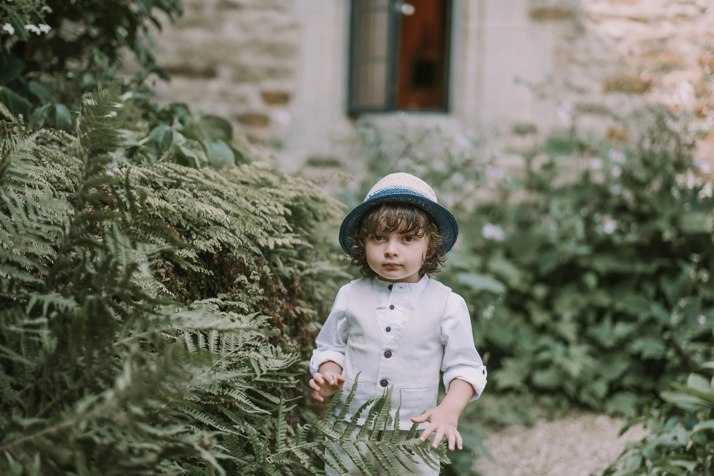 A young boy with curly hair, wearing a white shirt and a blue and gray hat, is standing outdoors among lush green ferns and foliage. There is a brick wall with a window in the background.