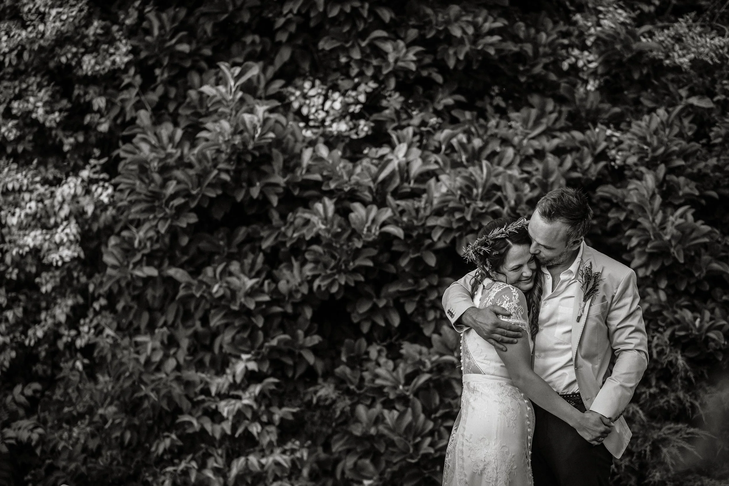 A black-and-white photo of a newlywed couple embracing outdoors, with lush foliage in the background.