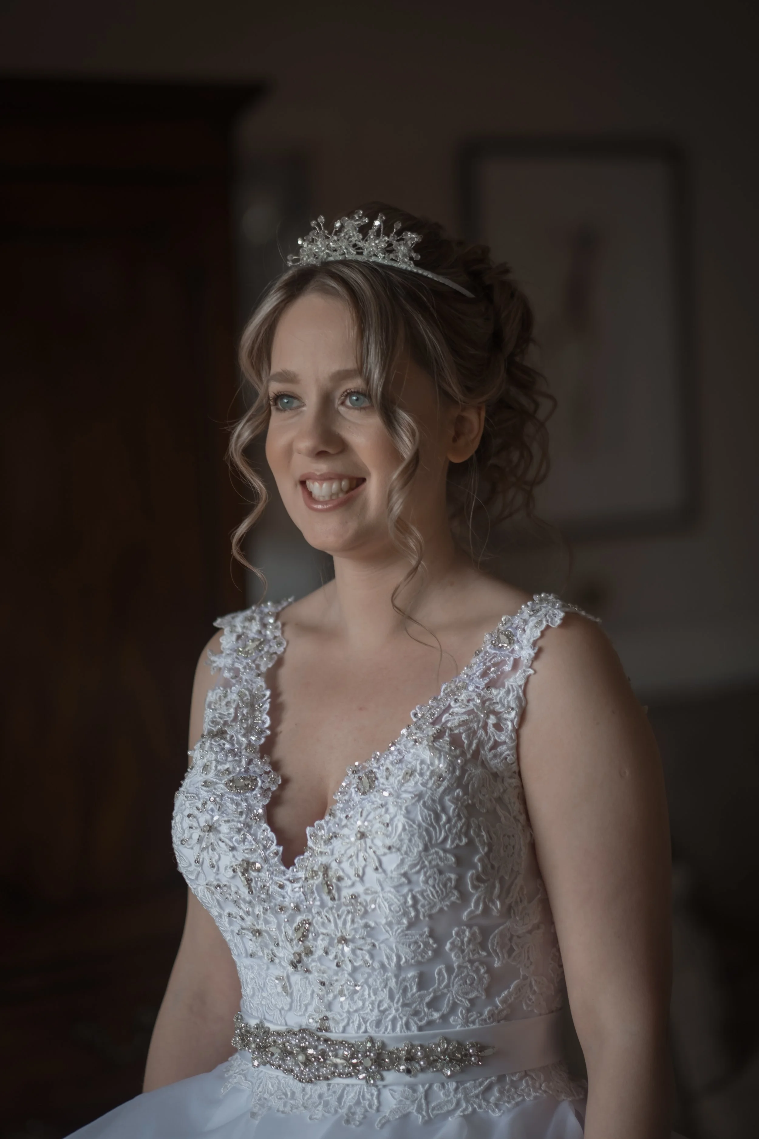 A smiling woman in a wedding dress with lace and bead details, wearing a tiara, and looking slightly to her right.