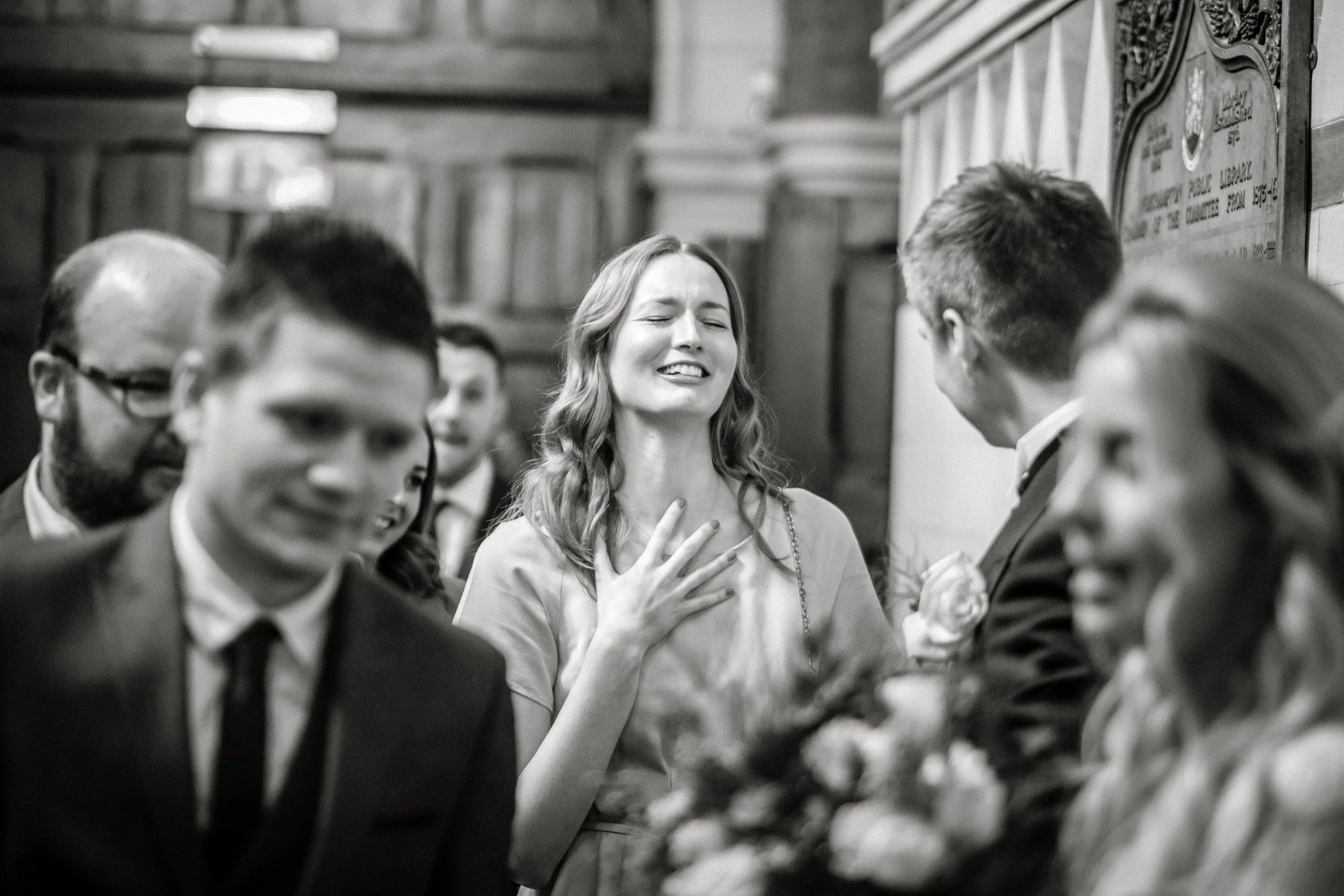 A woman with long hair and a delighted expression, standing in the center, with her hand over her chest, surrounded by formally dressed men, in what looks like a church or hall with wooden details.