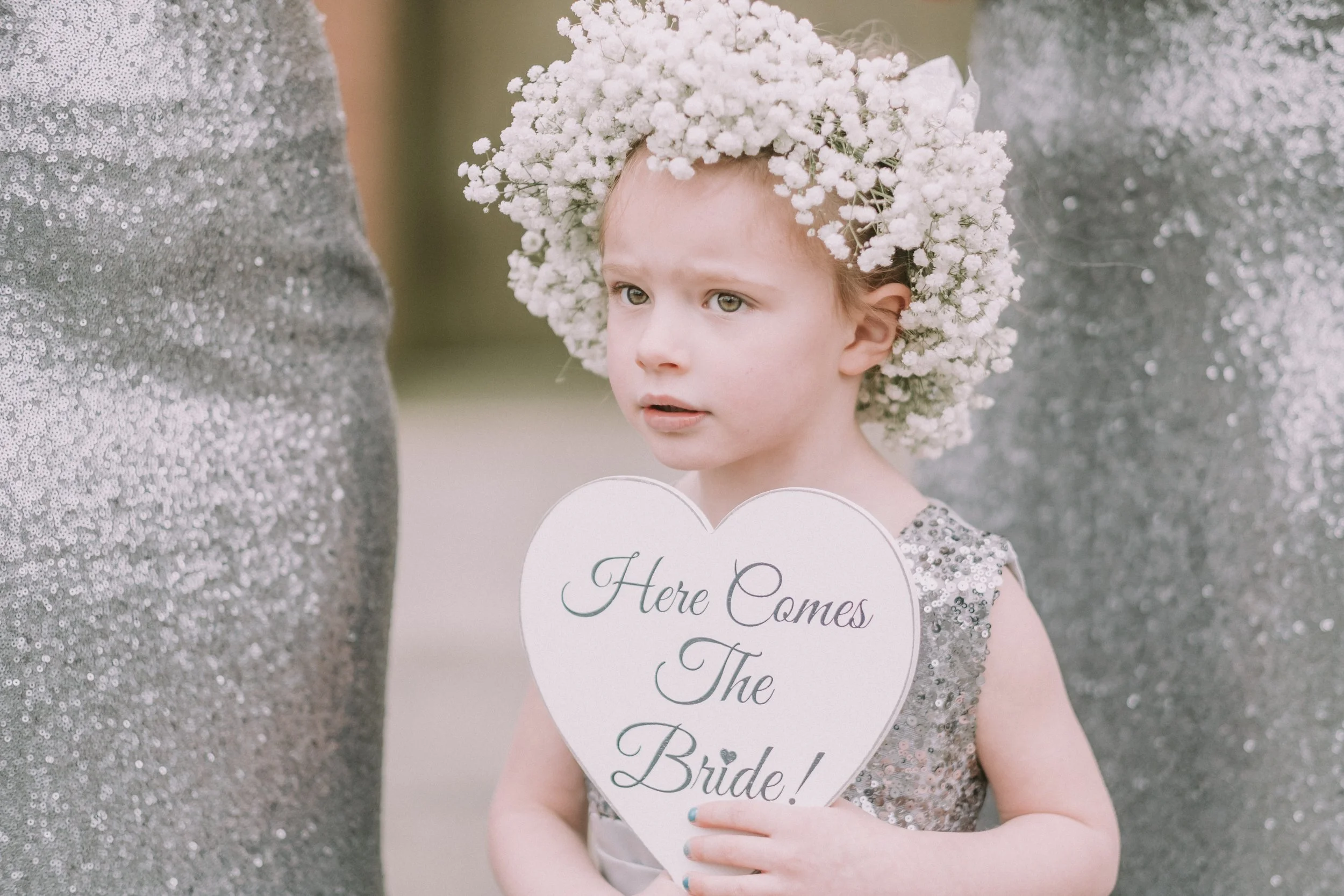 A young girl dressed in a sparkly dress wearing a large white floral crown, holding a heart-shaped sign that reads 'Here Comes The Bride!'