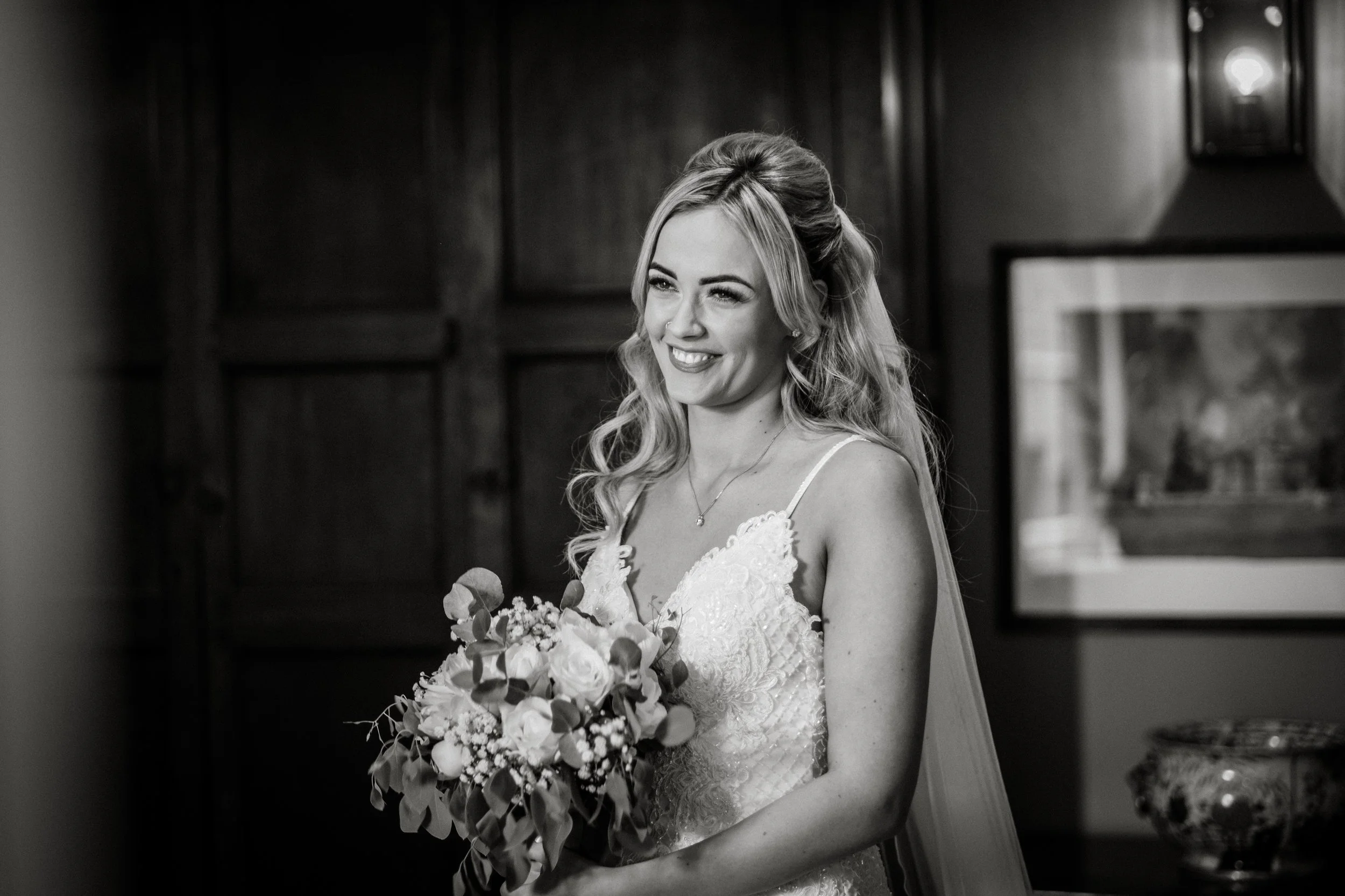 A smiling woman in a wedding dress holding a bouquet of flowers, standing indoors against a dark wooden wall.