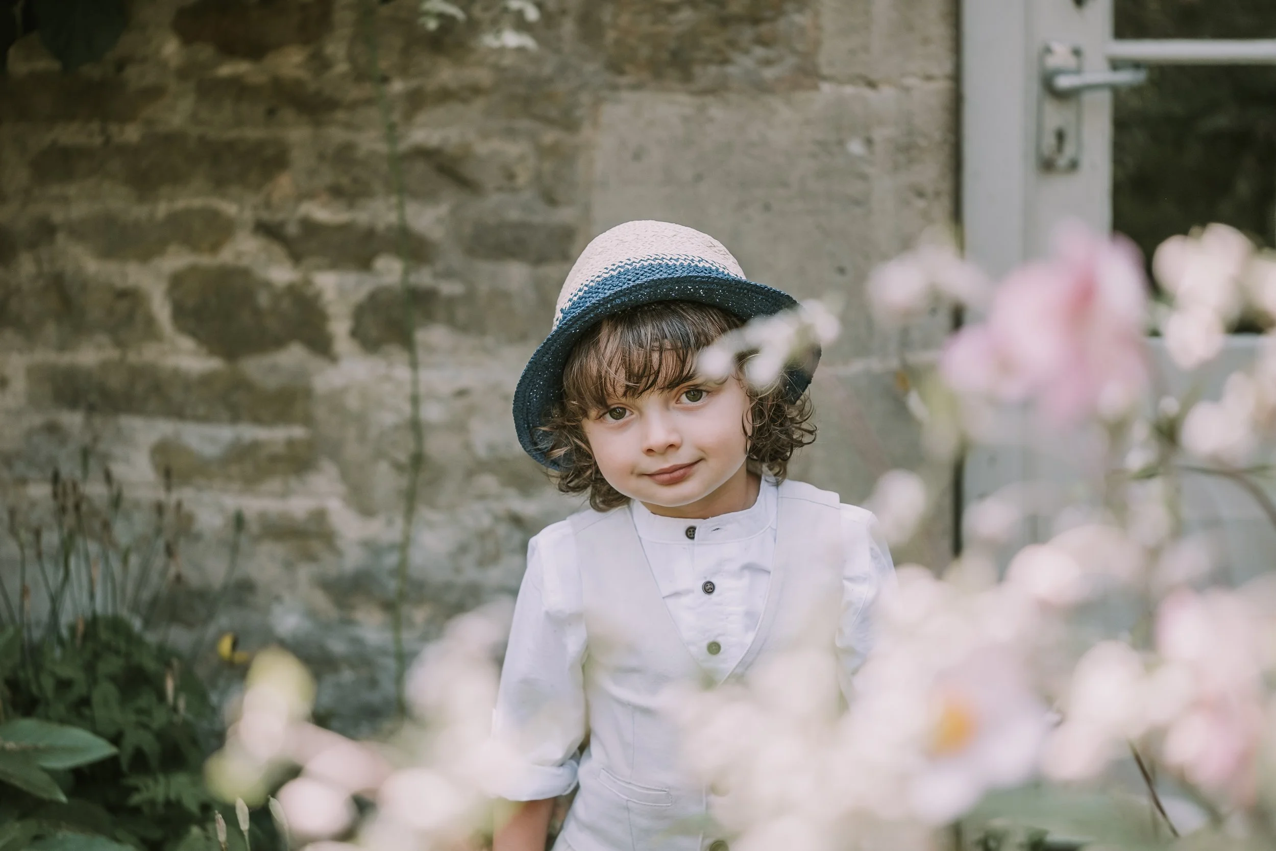 Young boy with curly hair wearing a white shirt and a straw hat standing among pink flowers in a garden.