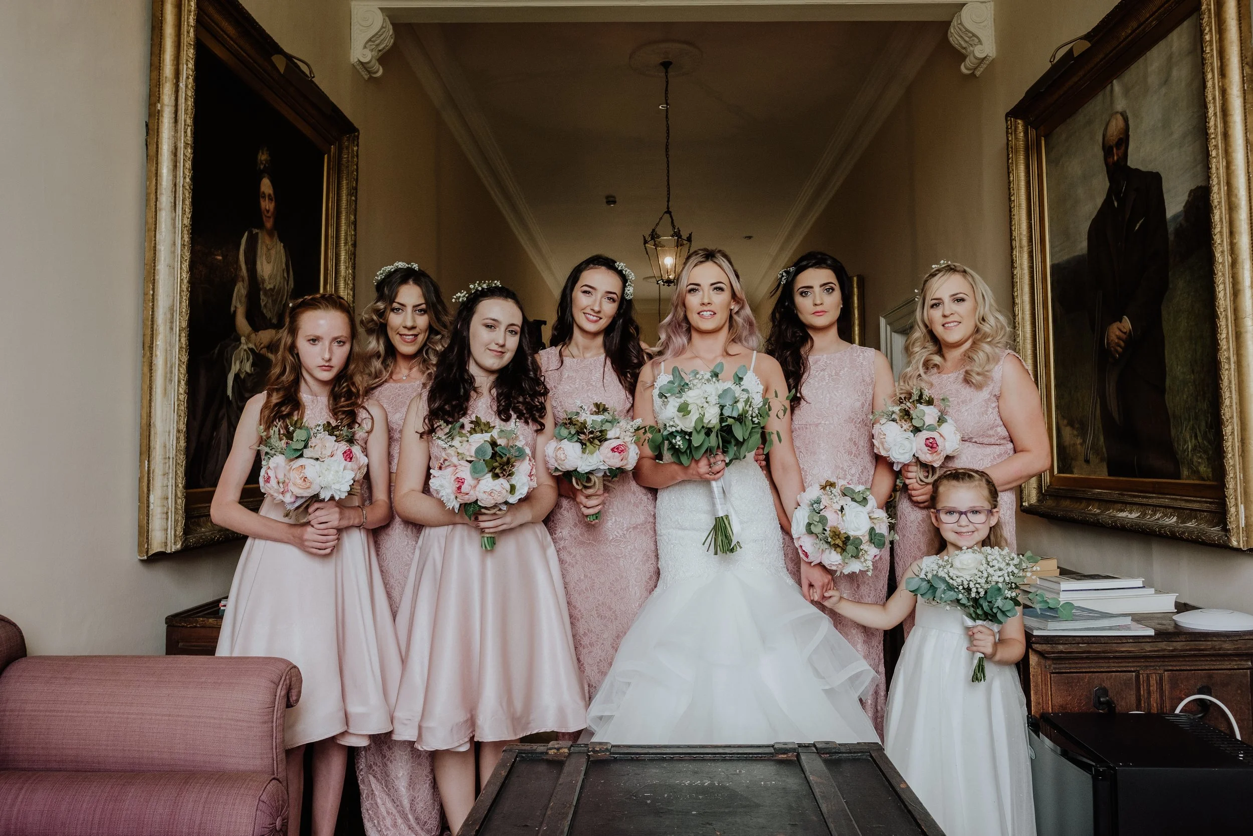 A group of seven women and two young girls, dressed in pink dresses and holding bouquets, pose for a photo at a wedding. The bride is in the center wearing a white gown, while the others are bridesmaids and flower girls standing around her. They are 