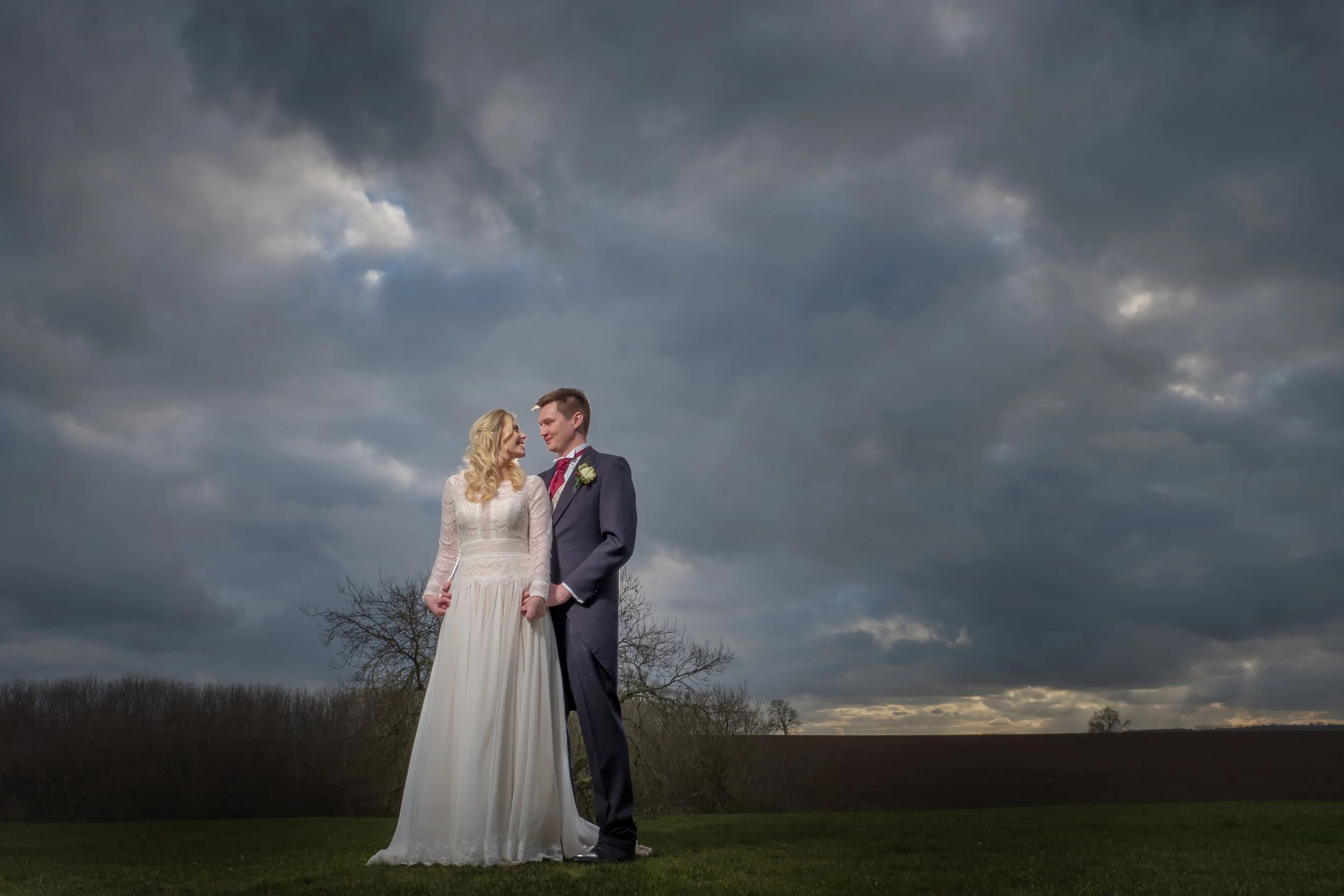 A bride and groom standing outdoors on a cloudy day, holding hands and looking at each other, with dark storm clouds overhead and a few bare trees in the background.