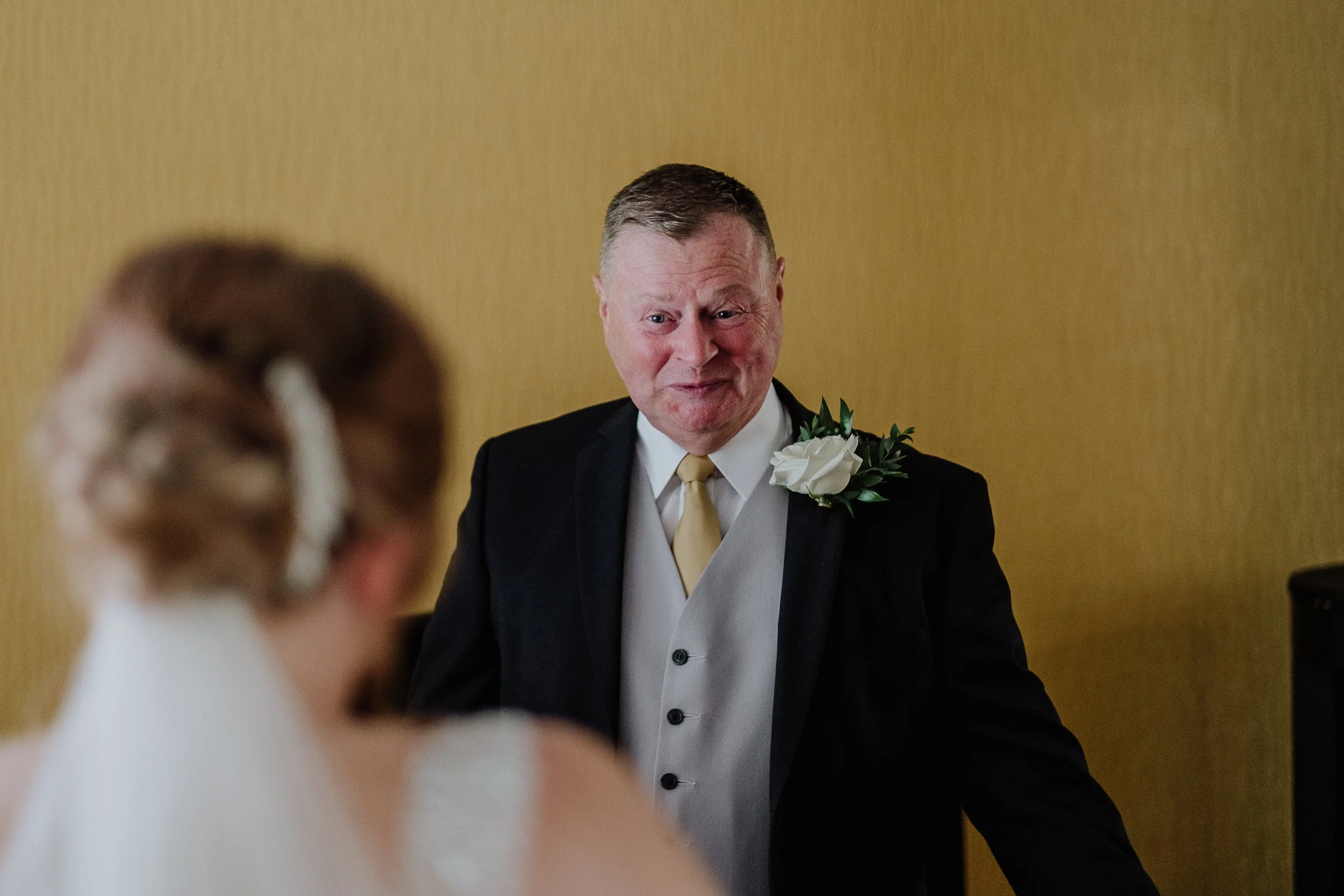 A middle-aged man with short brown hair, wearing a black suit, white shirt, yellow tie, and a white boutonniere, smiling and looking at a woman, in a formal setting with a plain yellow wall in the background.