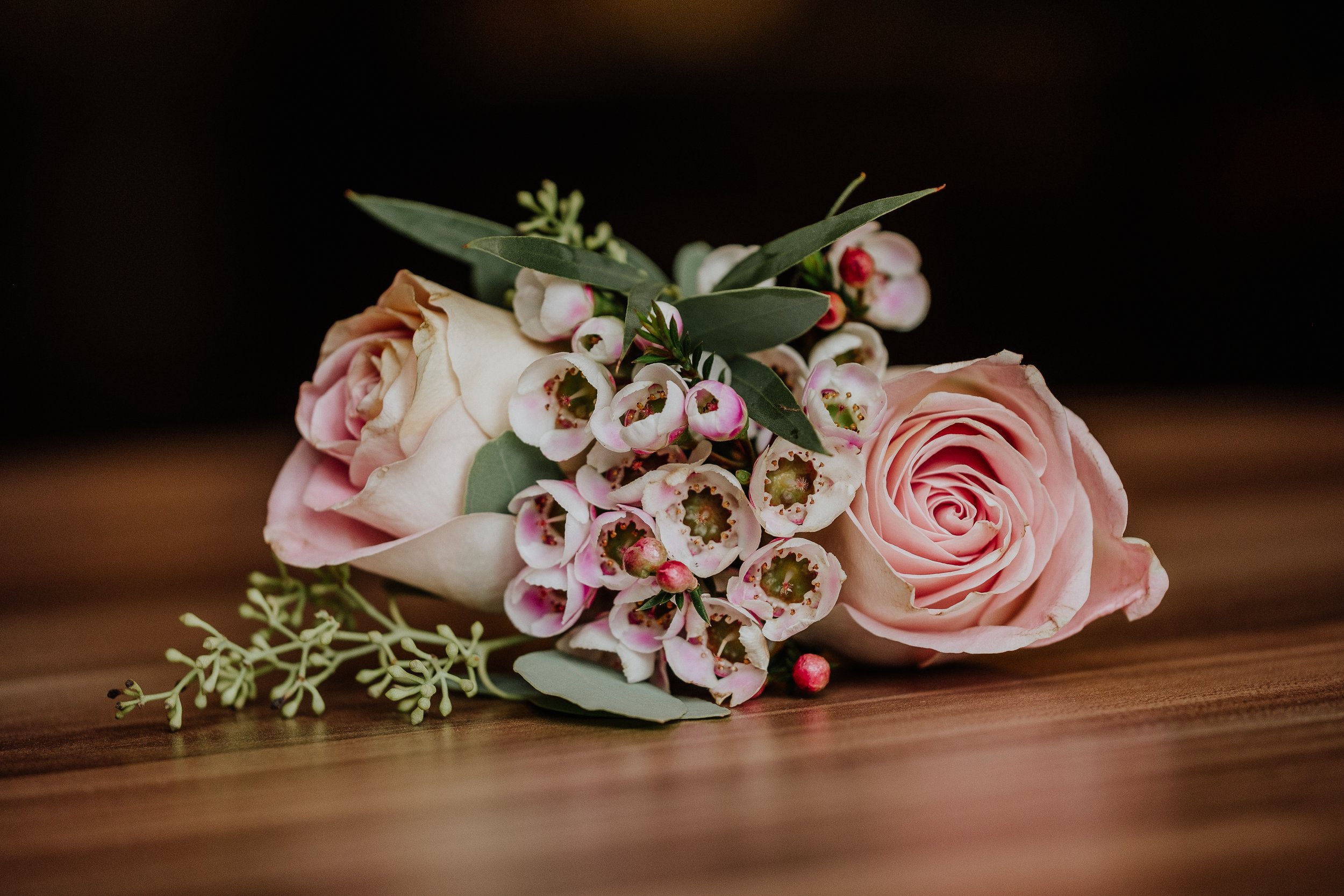 A bouquet of pale pink roses, white waxflowers with pink centers, greenery, and small white accents on a dark wooden surface.