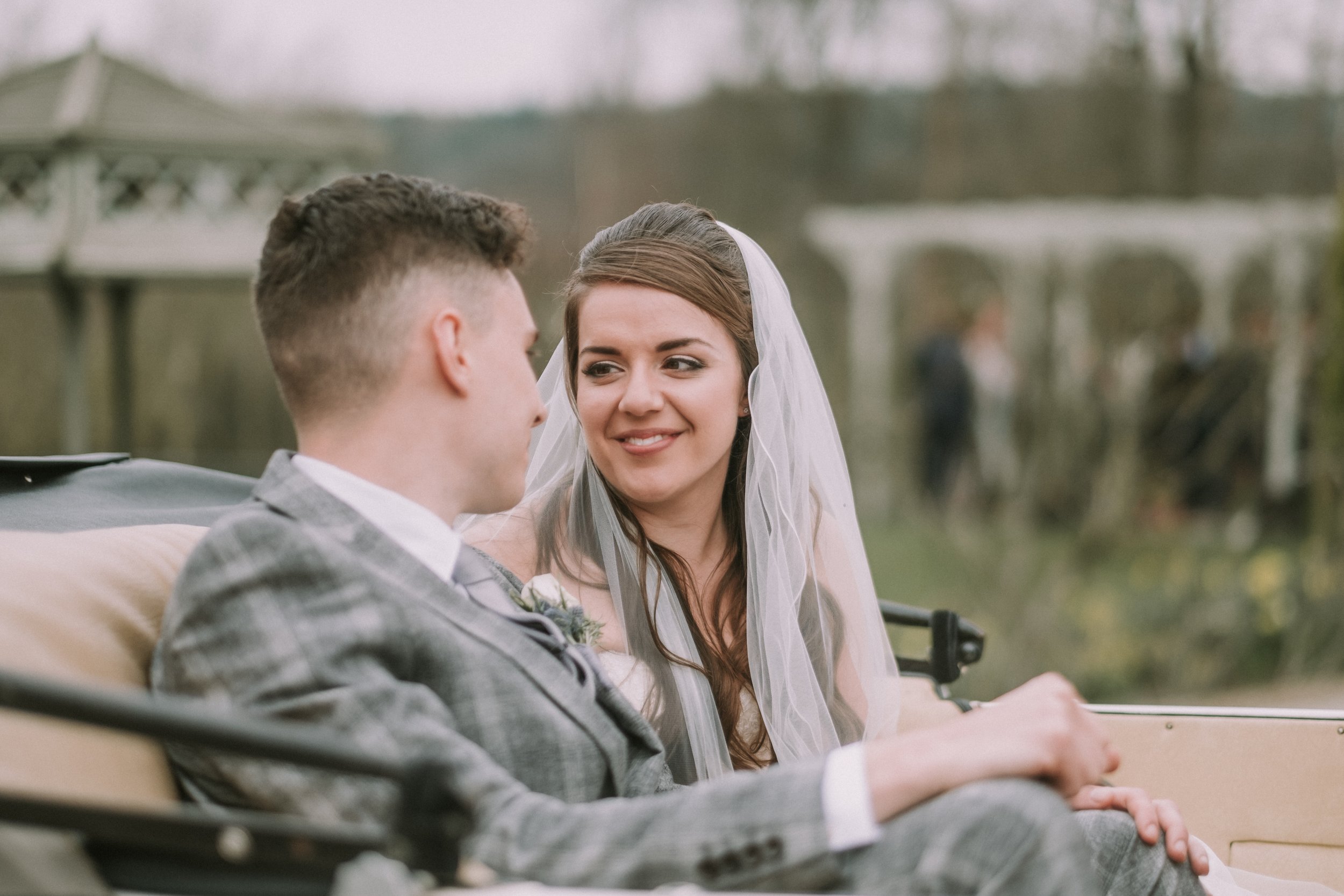 A bride and groom sit together on a vintage car during a wedding, outdoors with trees and a gazebo in the background.