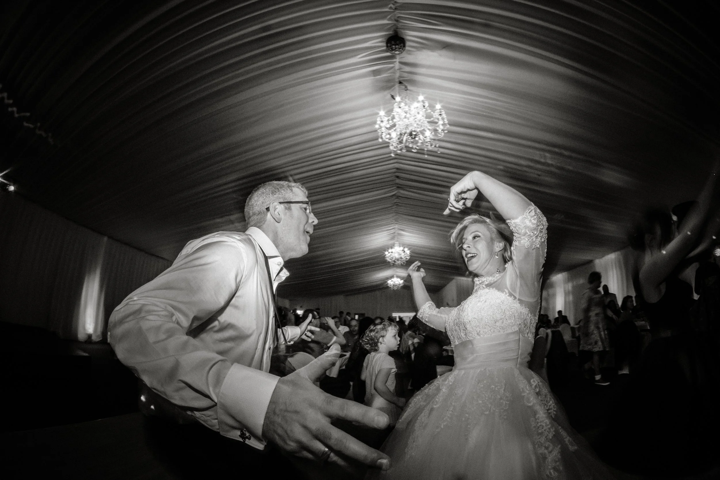 A couple dancing at a wedding reception, with the bride wearing a lace wedding dress and the groom in a dress shirt. The bride is smiling and lifting her arm while dancing. The venue has a tent ceiling with chandeliers, and guests can be seen in the 