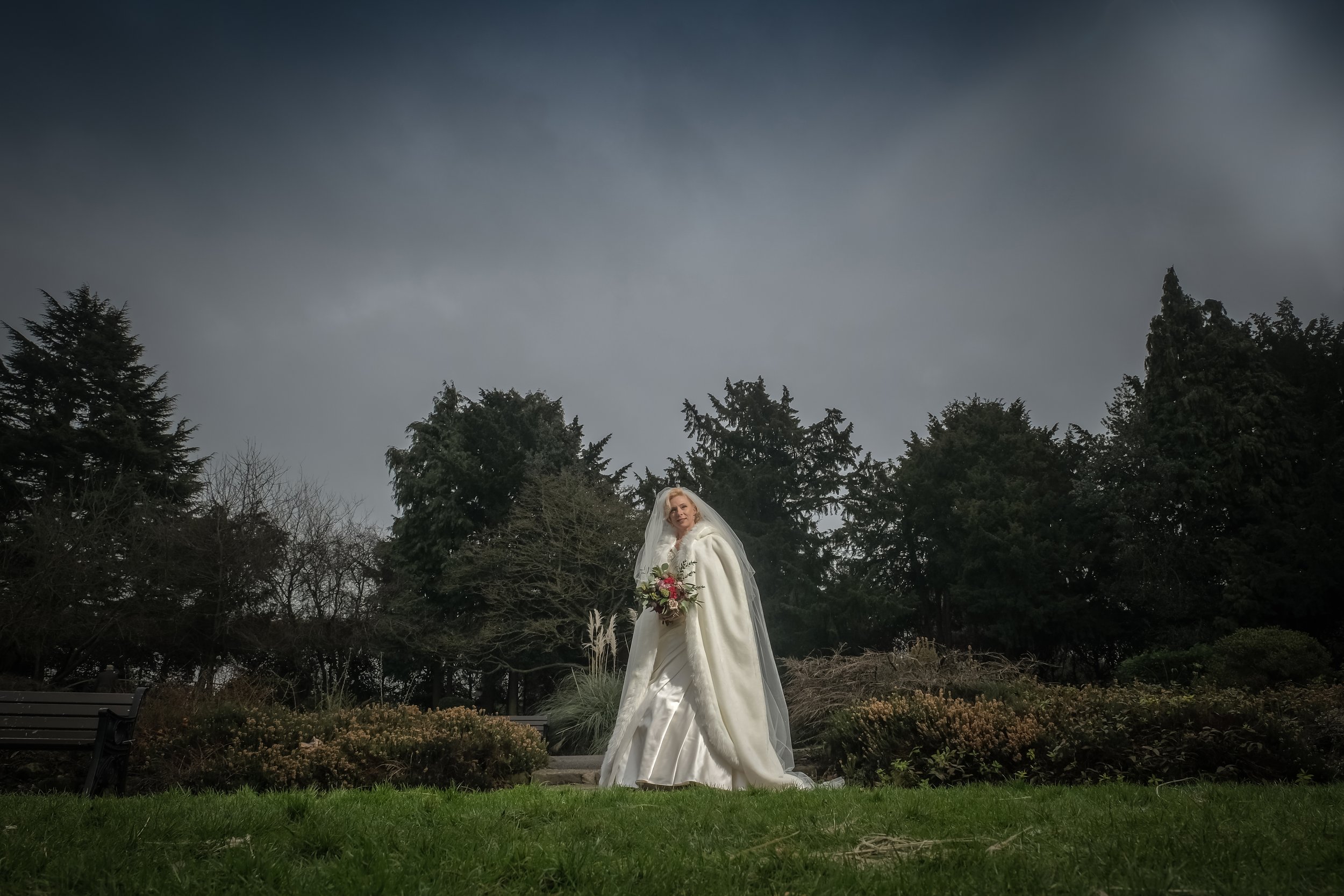A bride dressed in a white wedding gown and veil holding a bouquet standing outdoors near trees under a cloudy sky.