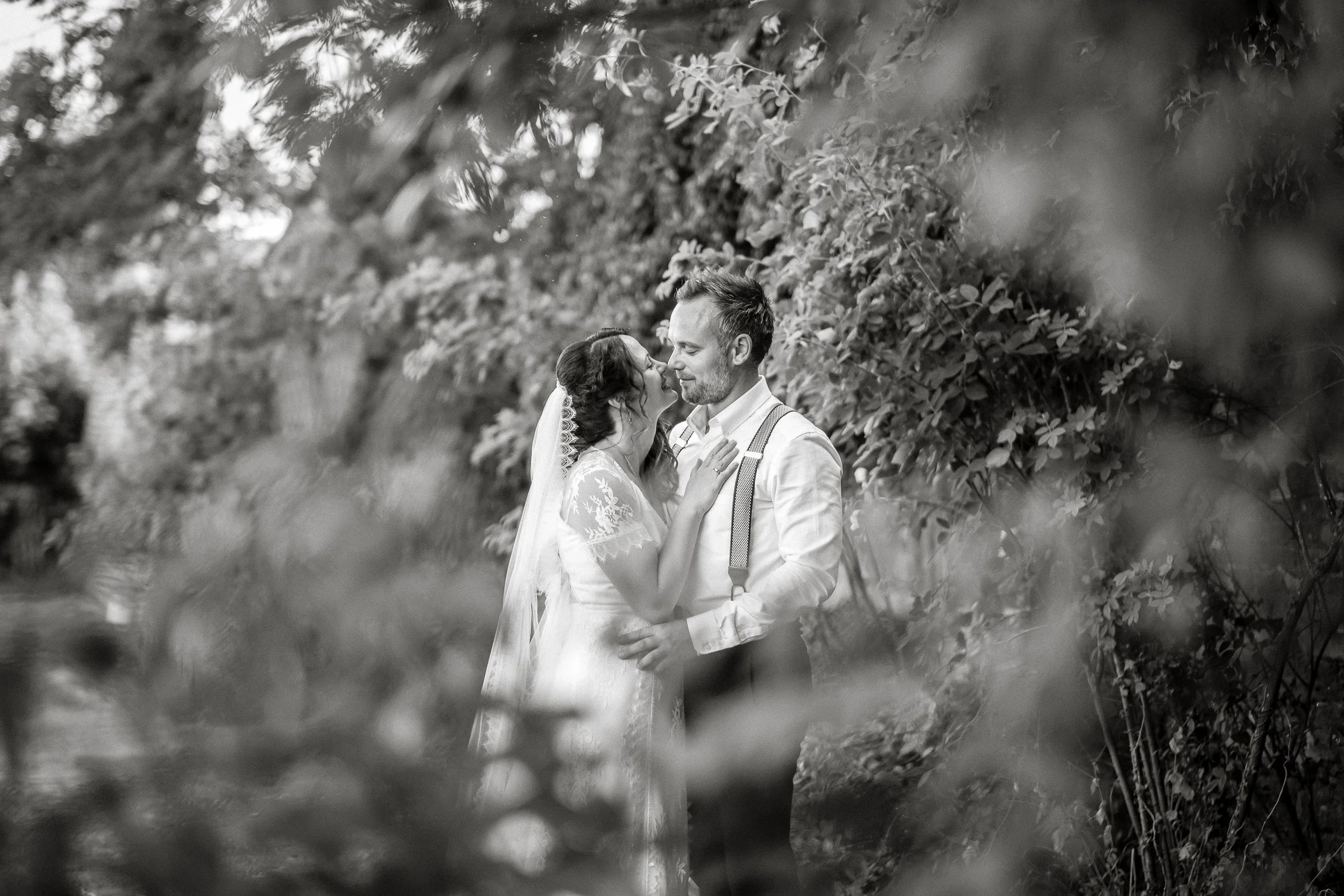 Black and white photo of a couple in wedding attire, standing close together and touching noses amidst lush foliage.