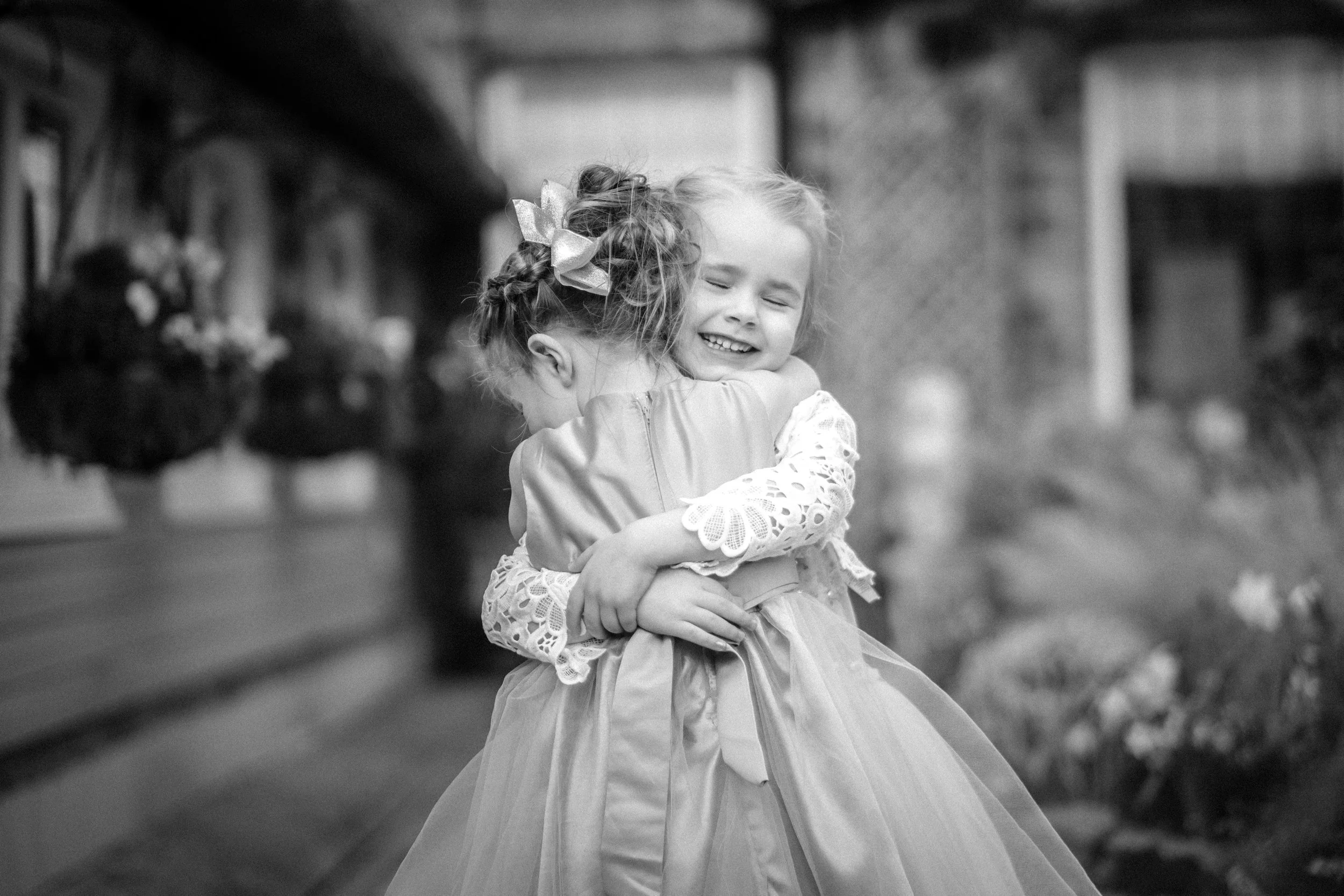 Two young girls in dresses embrace in a hug, with one girl smiling with eyes closed. The background suggests an outdoor setting with a building or structure.