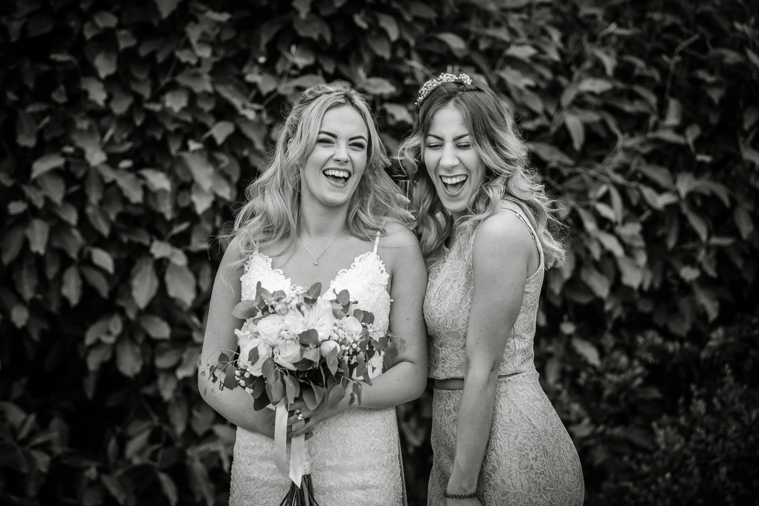 Two women, one holding a bouquet, are dressed in lace dresses and are laughing together outdoors against a backdrop of dense leaves.