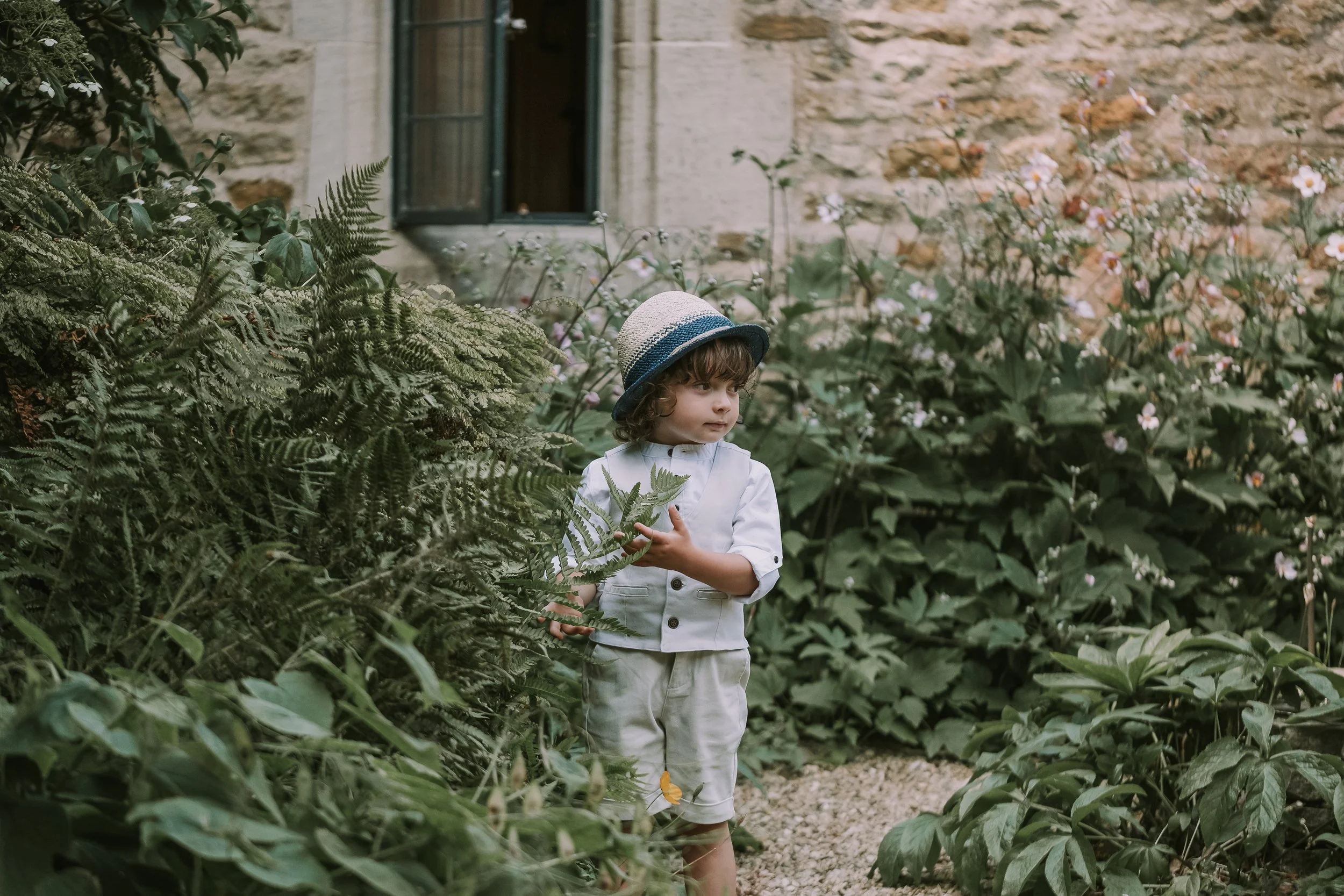 A young boy wearing a straw hat and white clothes standing among lush green plants and flowers outside a stone building with an open window.
