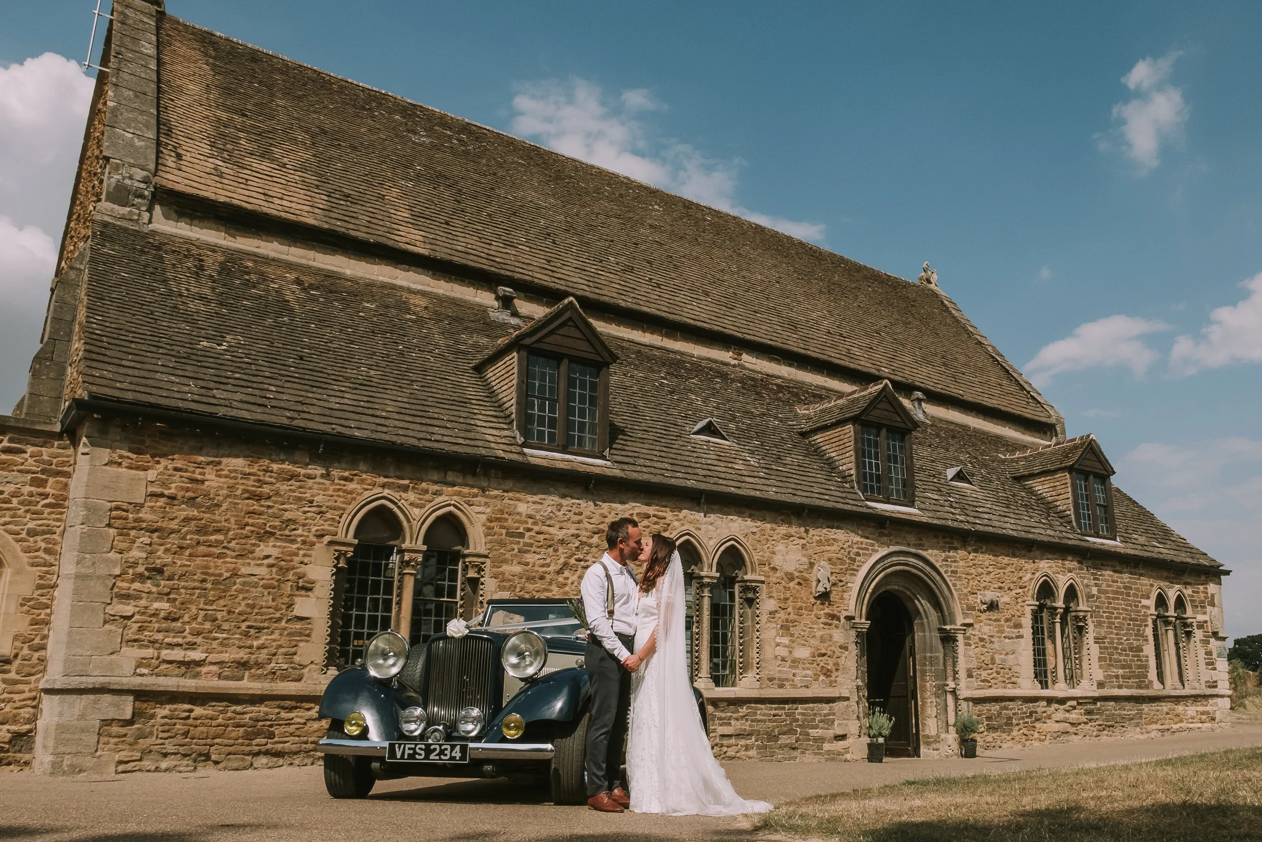 A couple dressed in wedding attire is standing in front of a vintage car outside a stone church, with the bride and groom gazing into each other's eyes, holding hands, under a partly cloudy sky.