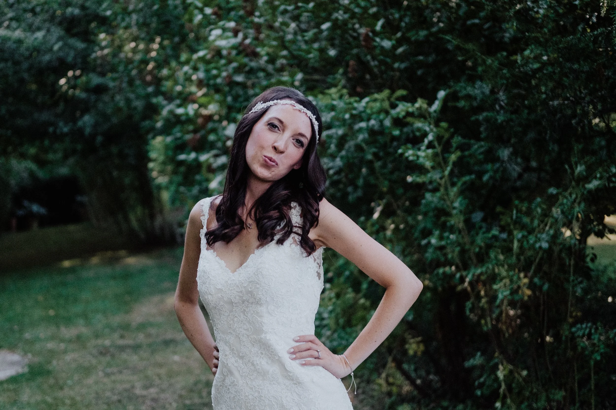 A woman in a white lace wedding dress posing outdoors with green trees in the background.