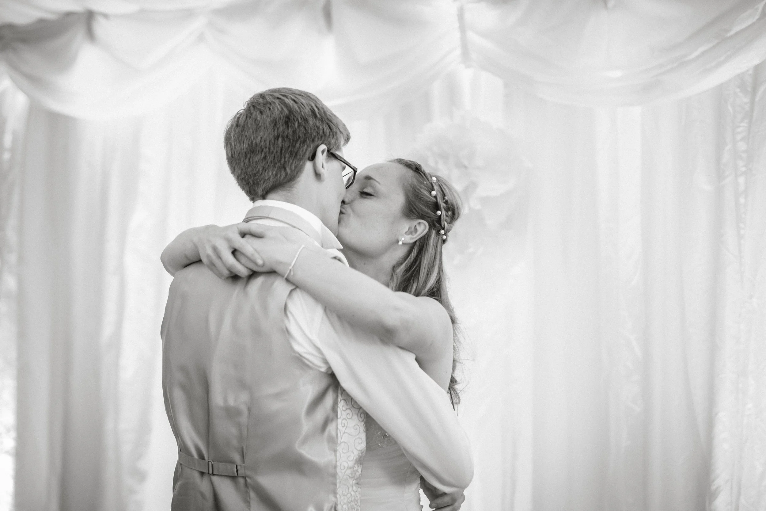 A black and white photo of a couple sharing a dance, the woman kissing the man while they embrace each other, with a decorated draped backdrop behind them.