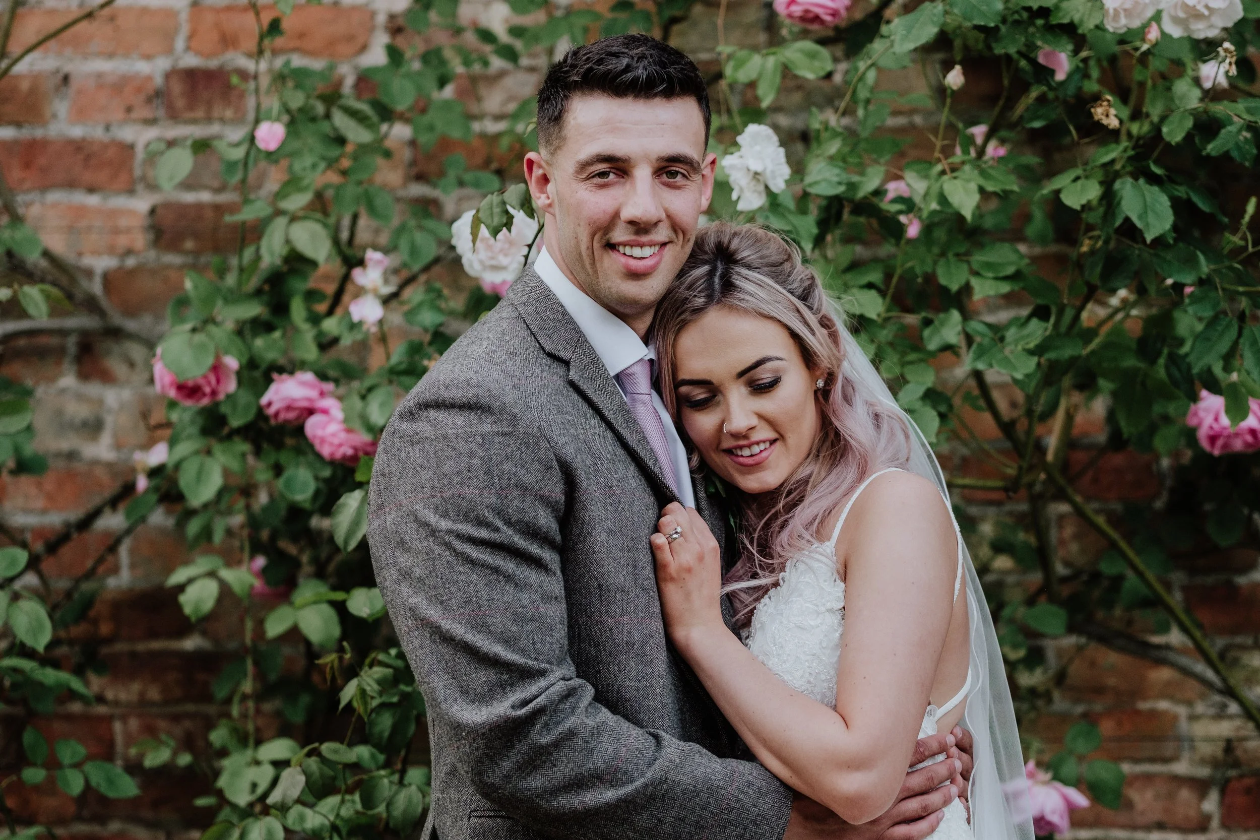 A happy couple, likely at their wedding, standing close together outdoors with a brick wall and blooming pink and white roses behind them. The groom is in a gray suit with a pink tie, and the bride has light hair with pink streaks, a veil, and a whit