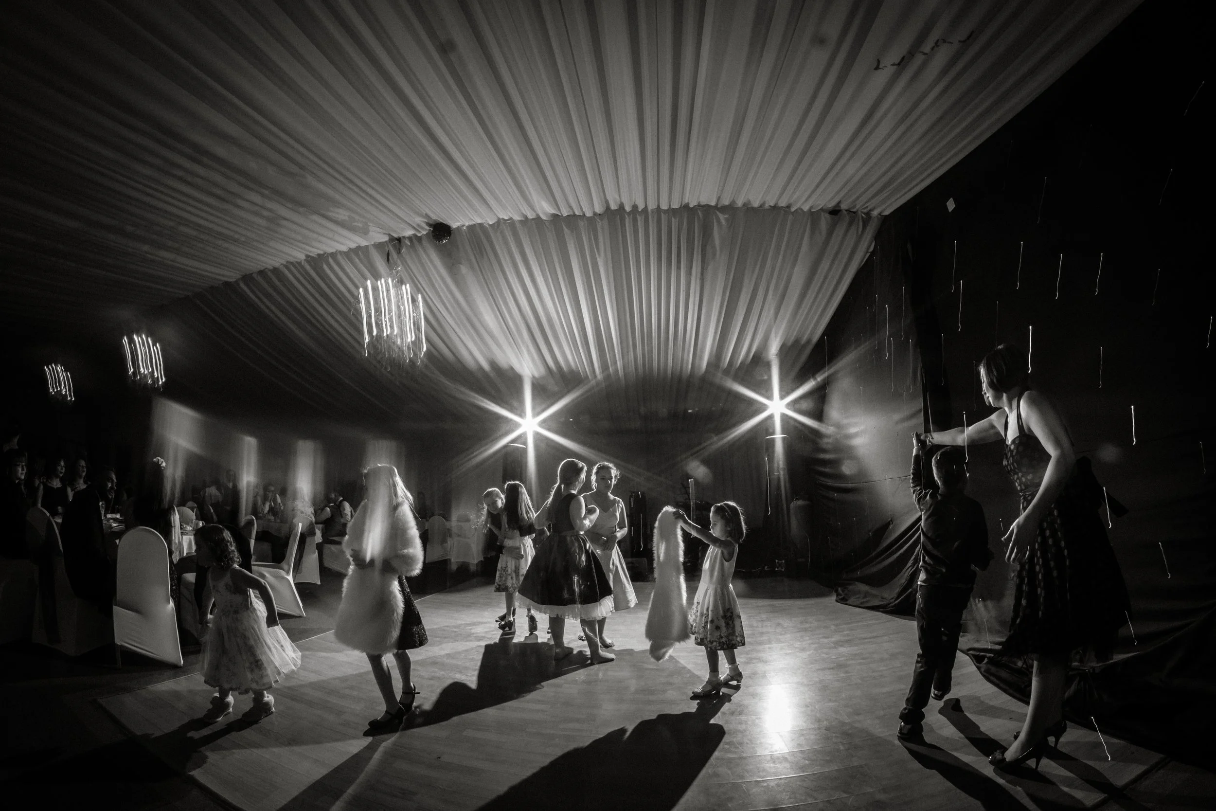 People at a party, some dancing and some sitting at tables, with dramatic lighting and a high ceiling decorated with fabric and hanging lights.