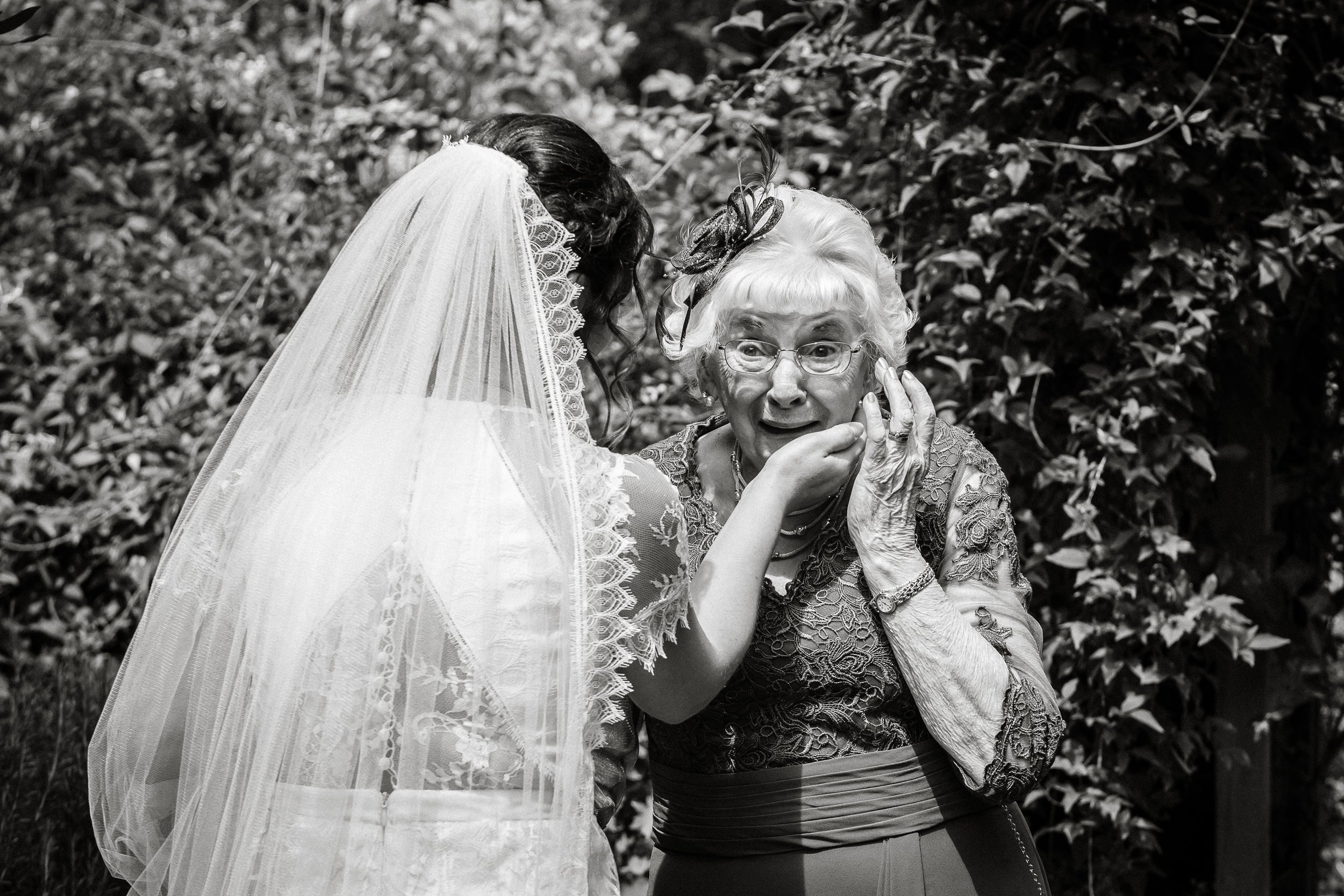 A bride in a wedding dress and veil leaning in to kiss an elderly woman with glasses and a lace blouse, outdoors with foliage in the background.