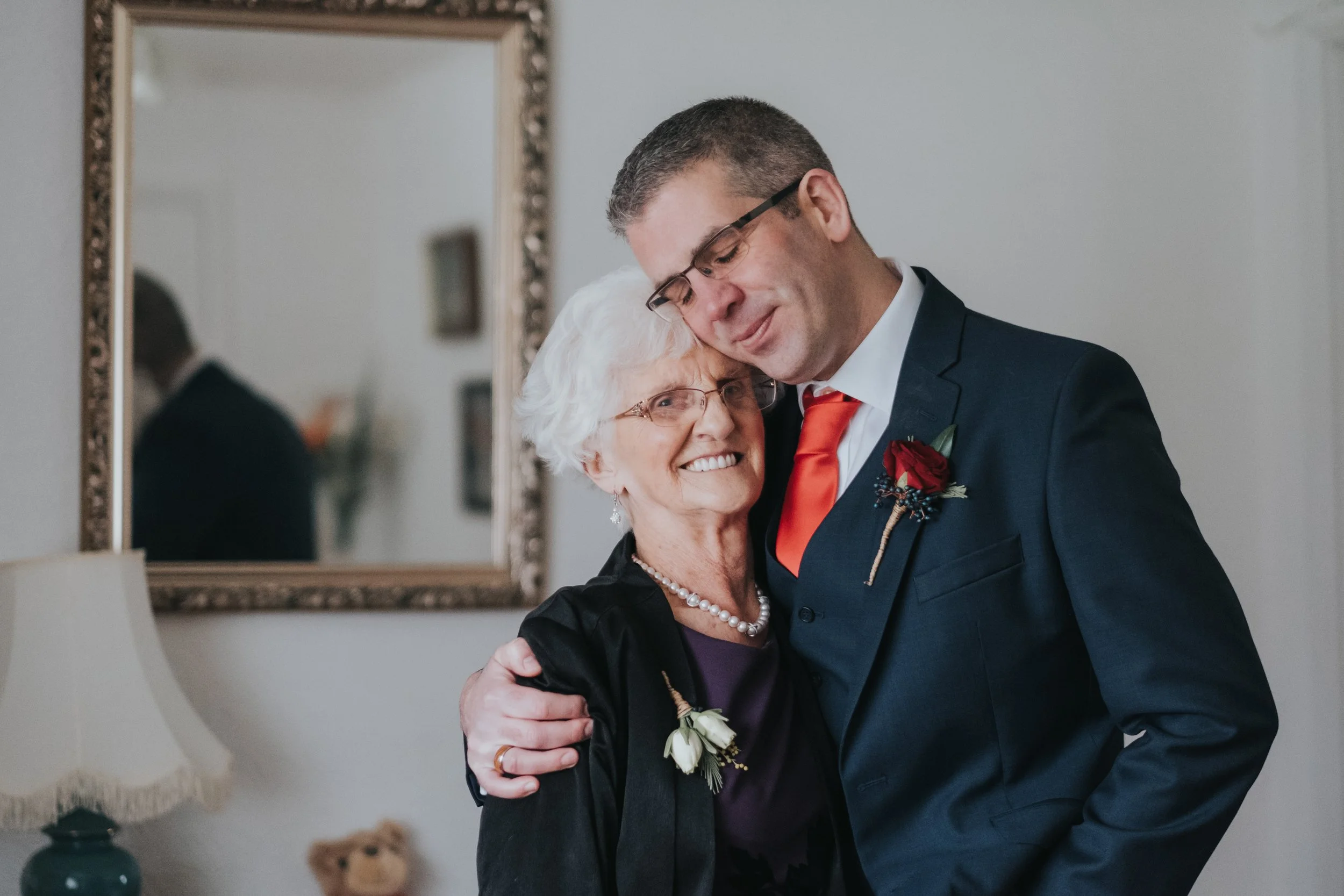 A young man and an elderly woman are hugging and smiling at each other in a warmly lit room, with a mirror, lamp, and blurred furniture in the background.