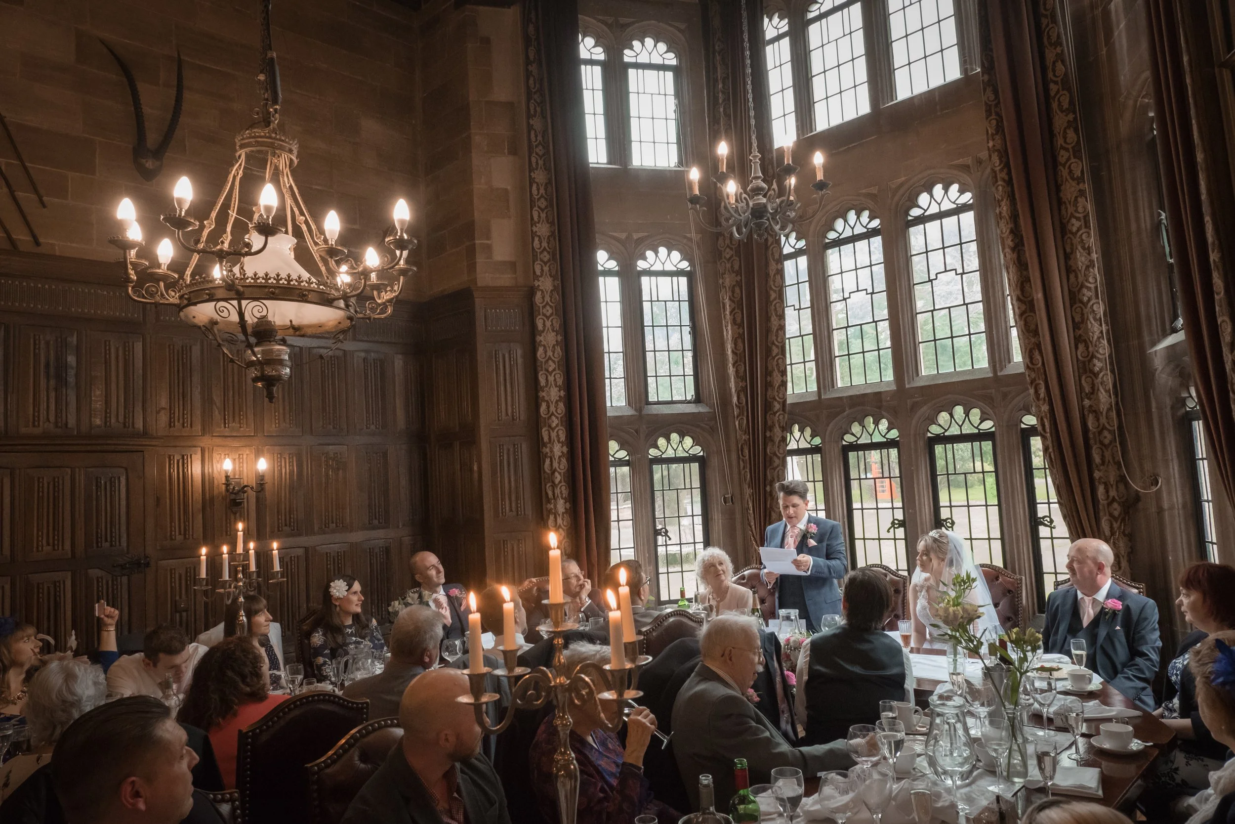 A wedding reception in an elegant, historic room with large ornate windows, wooden paneling, and chandeliers. People are seated at long tables with candles and floral arrangements, while a man in a suit is giving a speech.